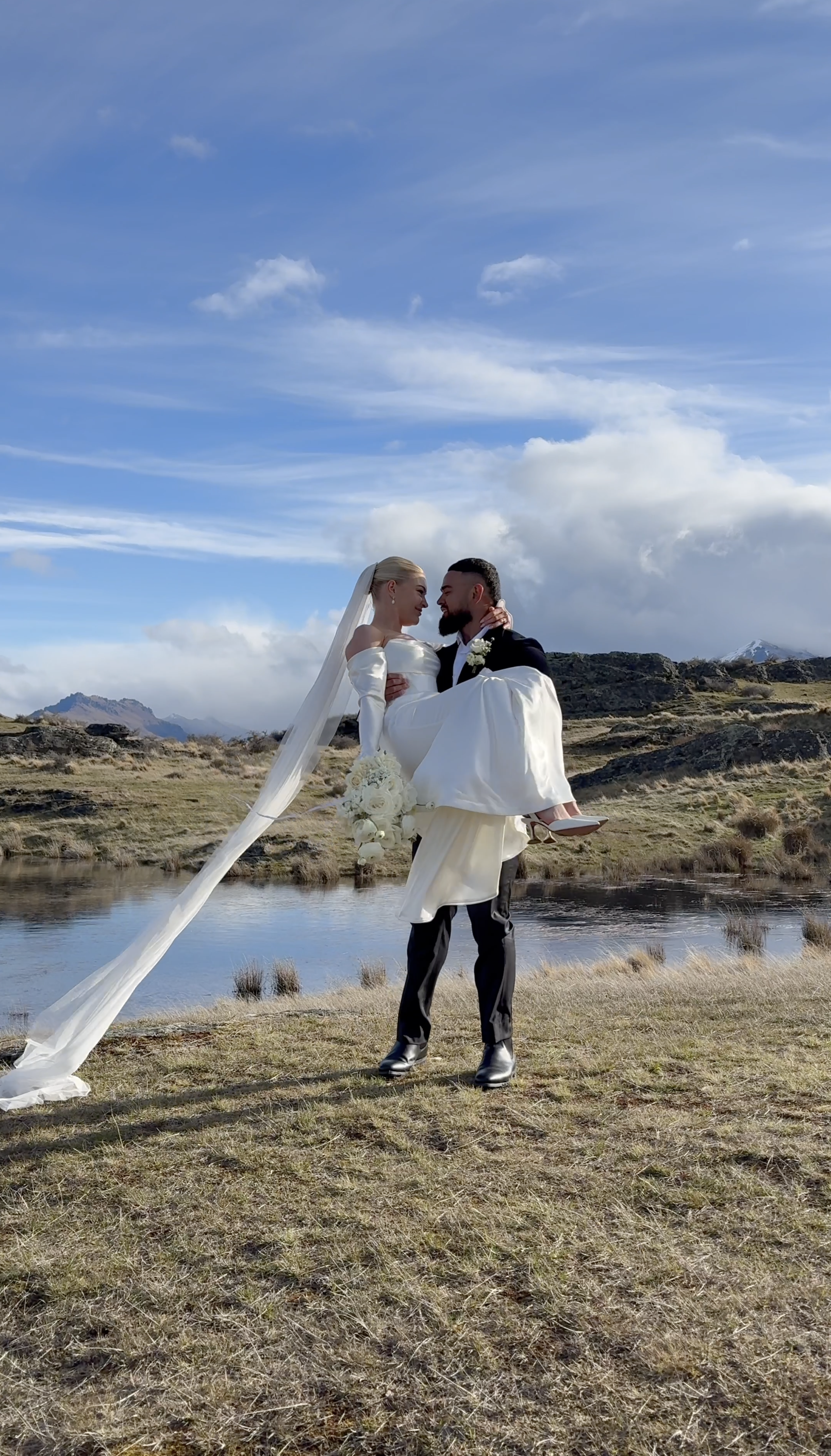 A bride and groom share a romantic moment outdoors near a small lake, with the groom holding the bride in his arms. The bride is wearing a long-sleeved white wedding dress with a veil, while the groom is dressed in a dark suit. The landscape features