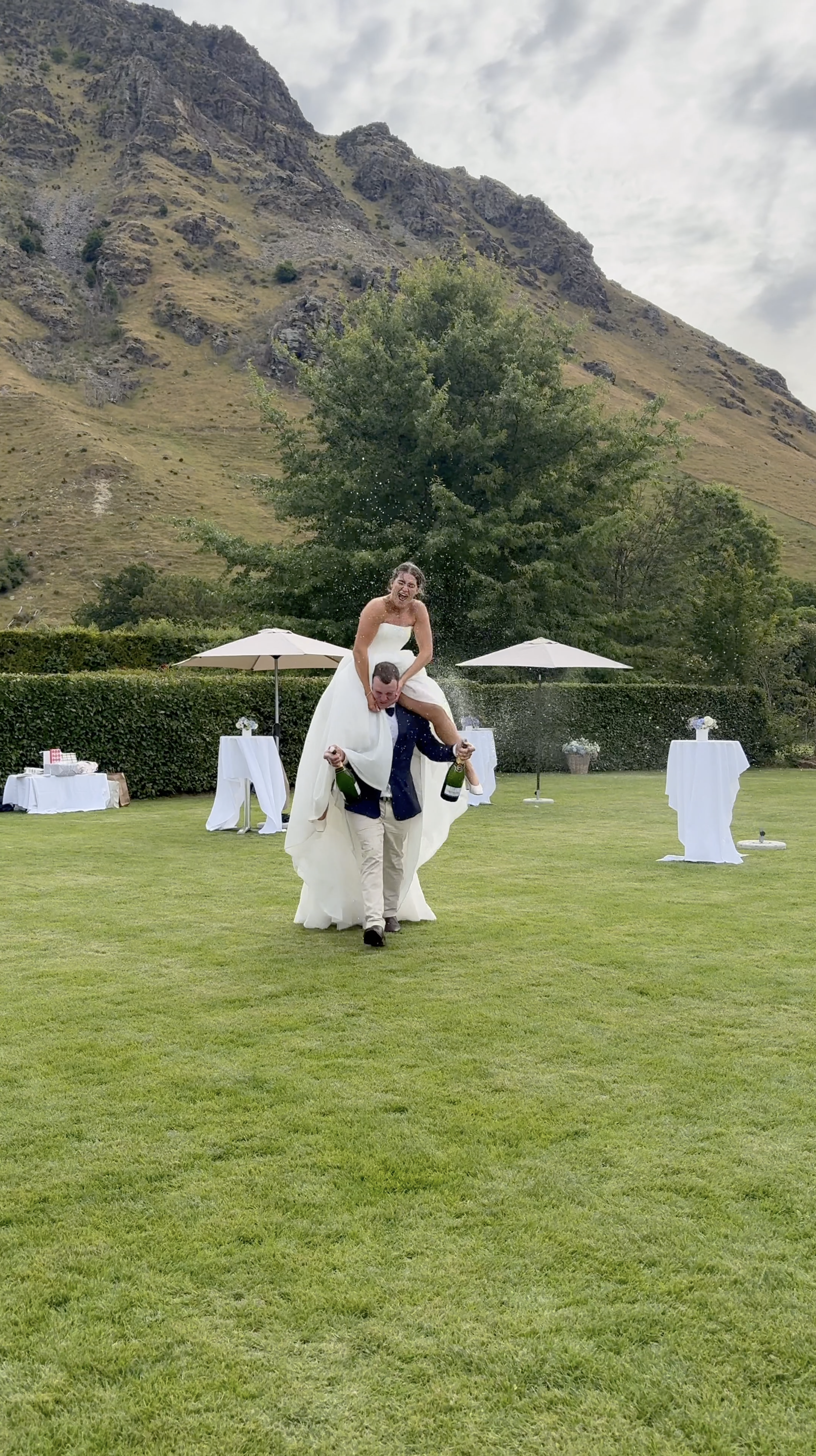 A bride and groom celebrating outdoors at a wedding, with the groom carrying the bride on his shoulders, both holding champagne bottles, on a grassy lawn with mountains and trees in the background. Captured by a wedding content creator 