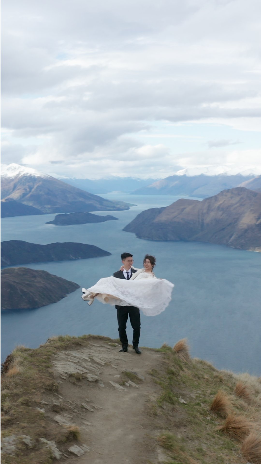 A couple in wedding attire, with the groom in a suit and the bride in a wedding dress, standing on a mountain ledge overlooking a lake and mountain ranges, the groom lifting the bride in his arms.