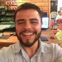 Gülen genç bir adam, arka planda bir kafe veya restoran ortamı.