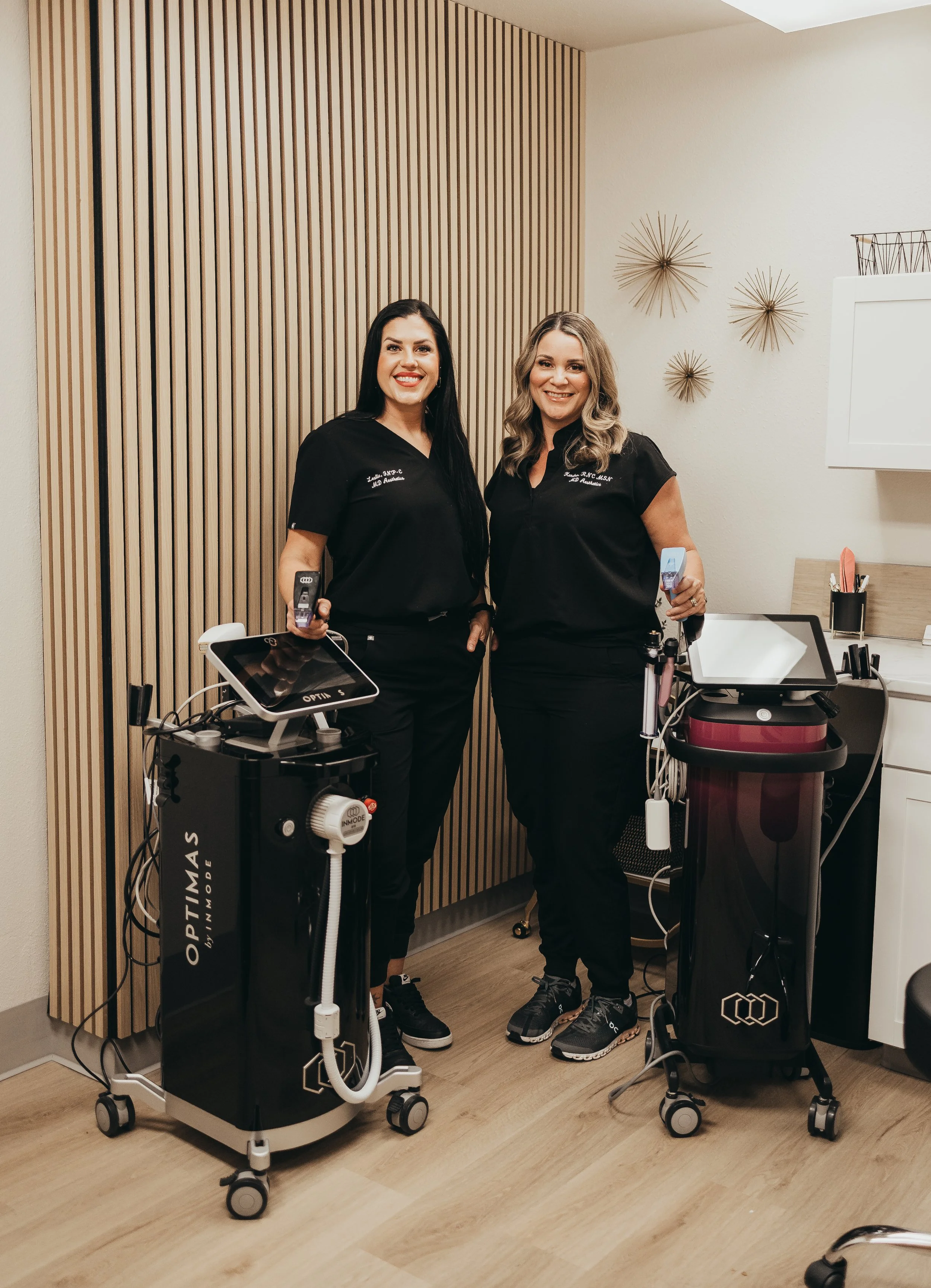 Two women in medical scrubs standing in a medical office, smiling, with medical equipment around them.