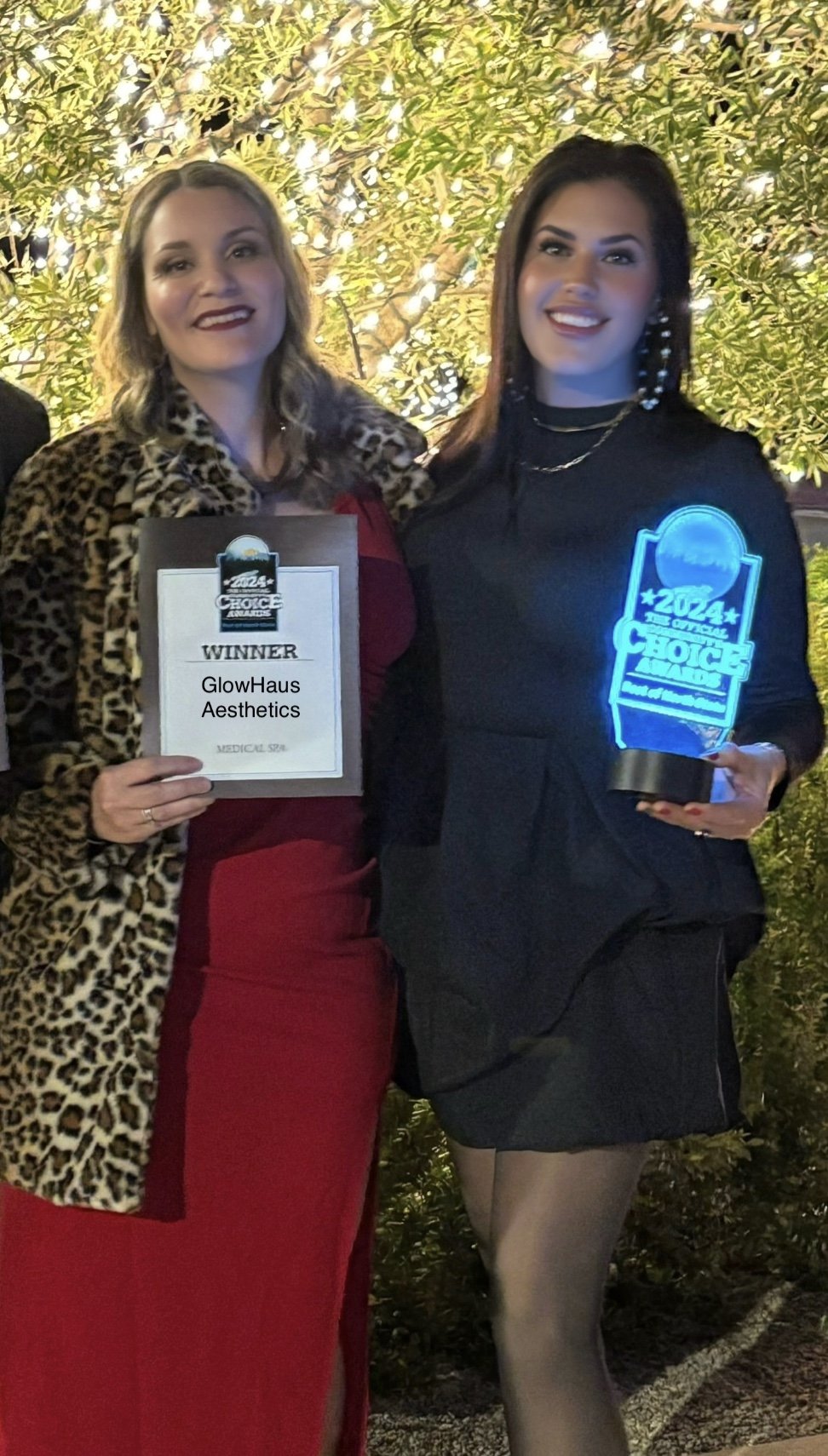 Two women standing outdoors at night, celebrating an award. The woman on the left wears a red dress and a leopard print jacket, holding a certificate that reads 'Winner GlowHaus Aesthetics.' The woman on the right wears a black outfit and holds a glo