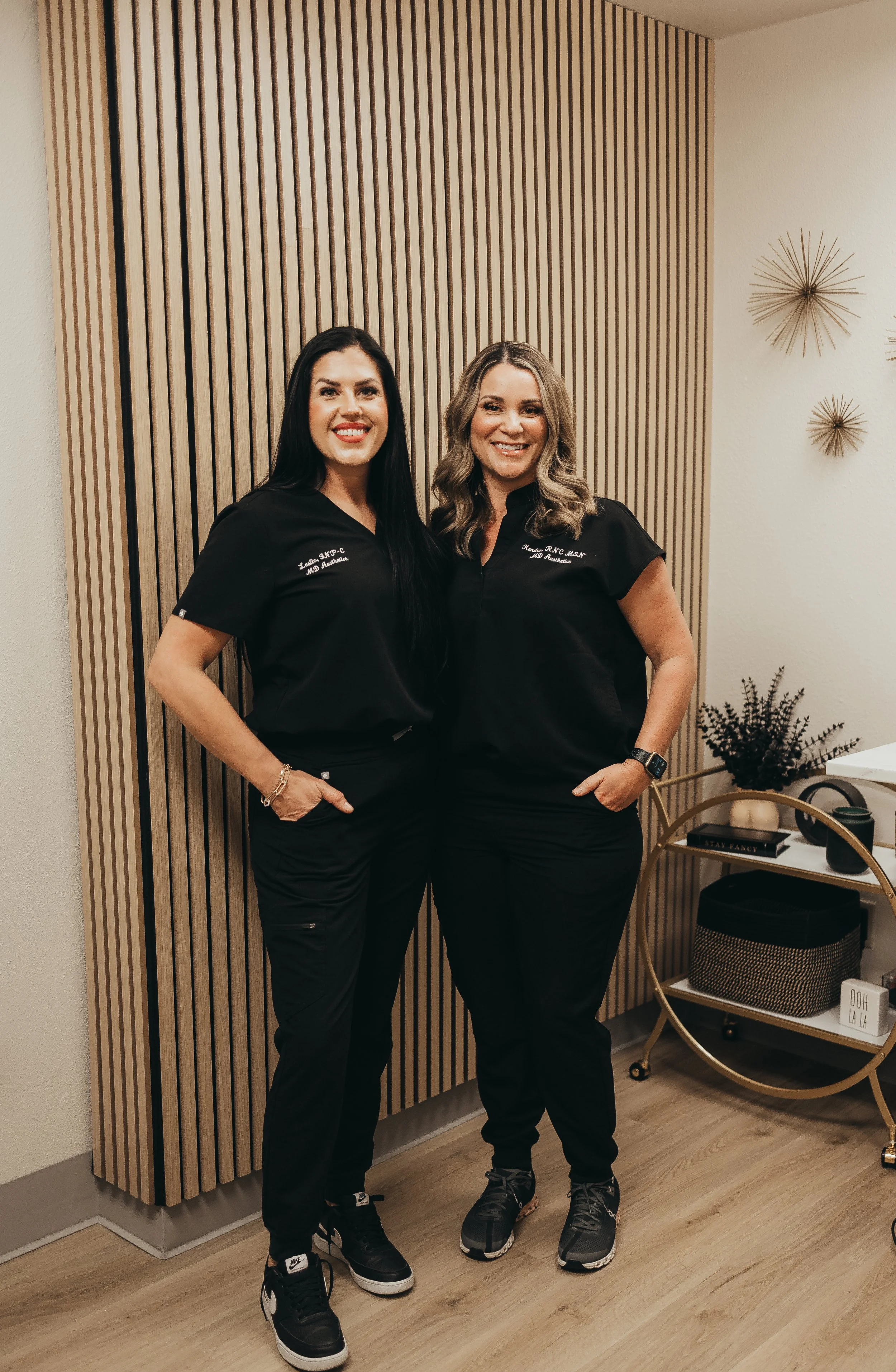 Two women in black scrubs standing inside a room with a wooden slat wall behind them, smiling at the camera.
