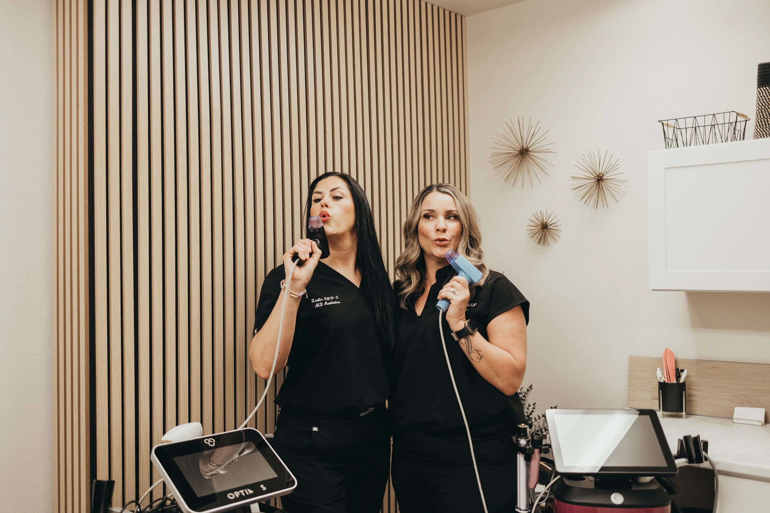 Two women in medical scrubs with hair rollers, holding electronic devices, standing in an office or clinic setting.