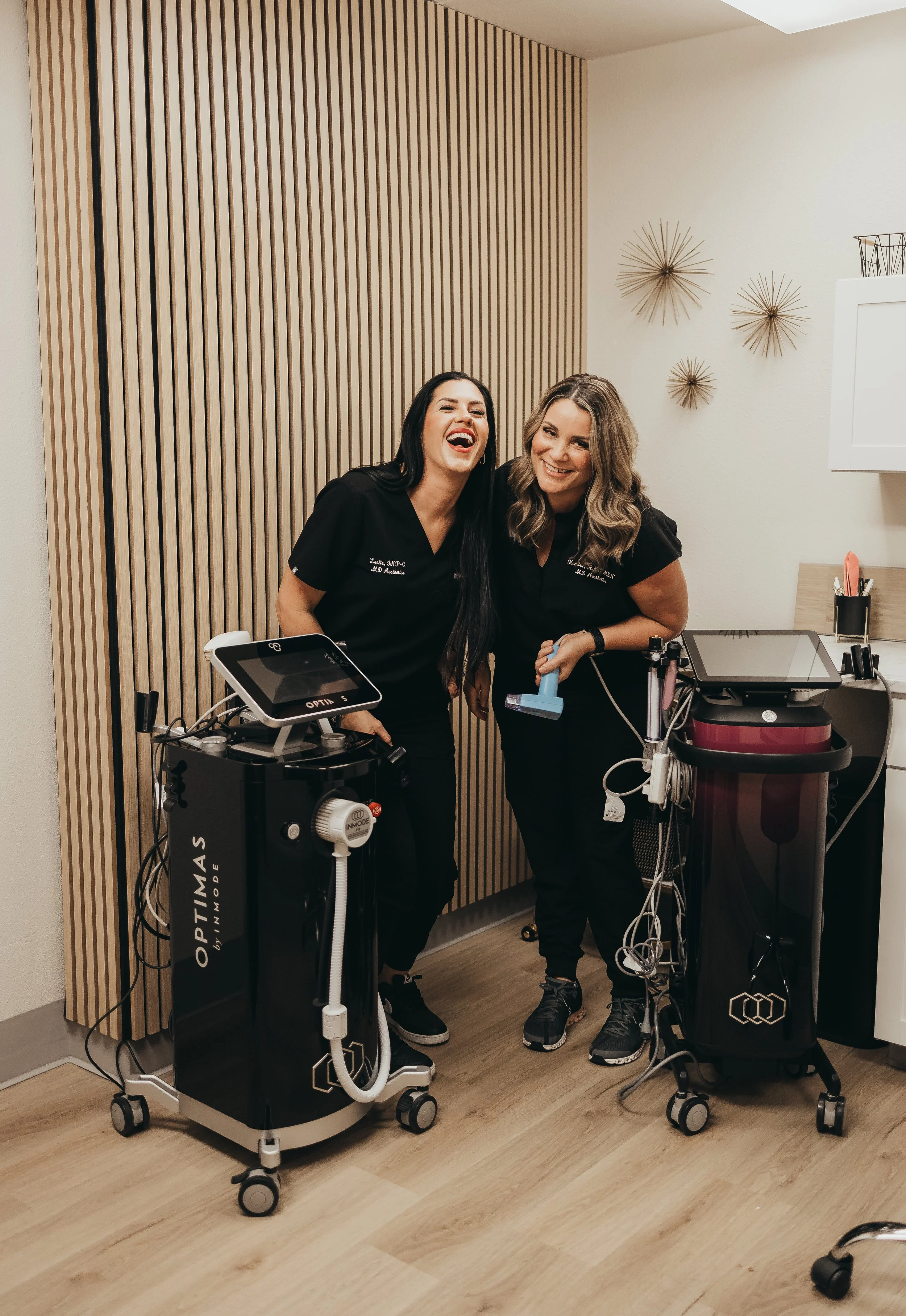 Two women in black medical scrubs smiling and posing in a medical office, surrounded by medical equipment including a mobile device, a machine, and cables.
