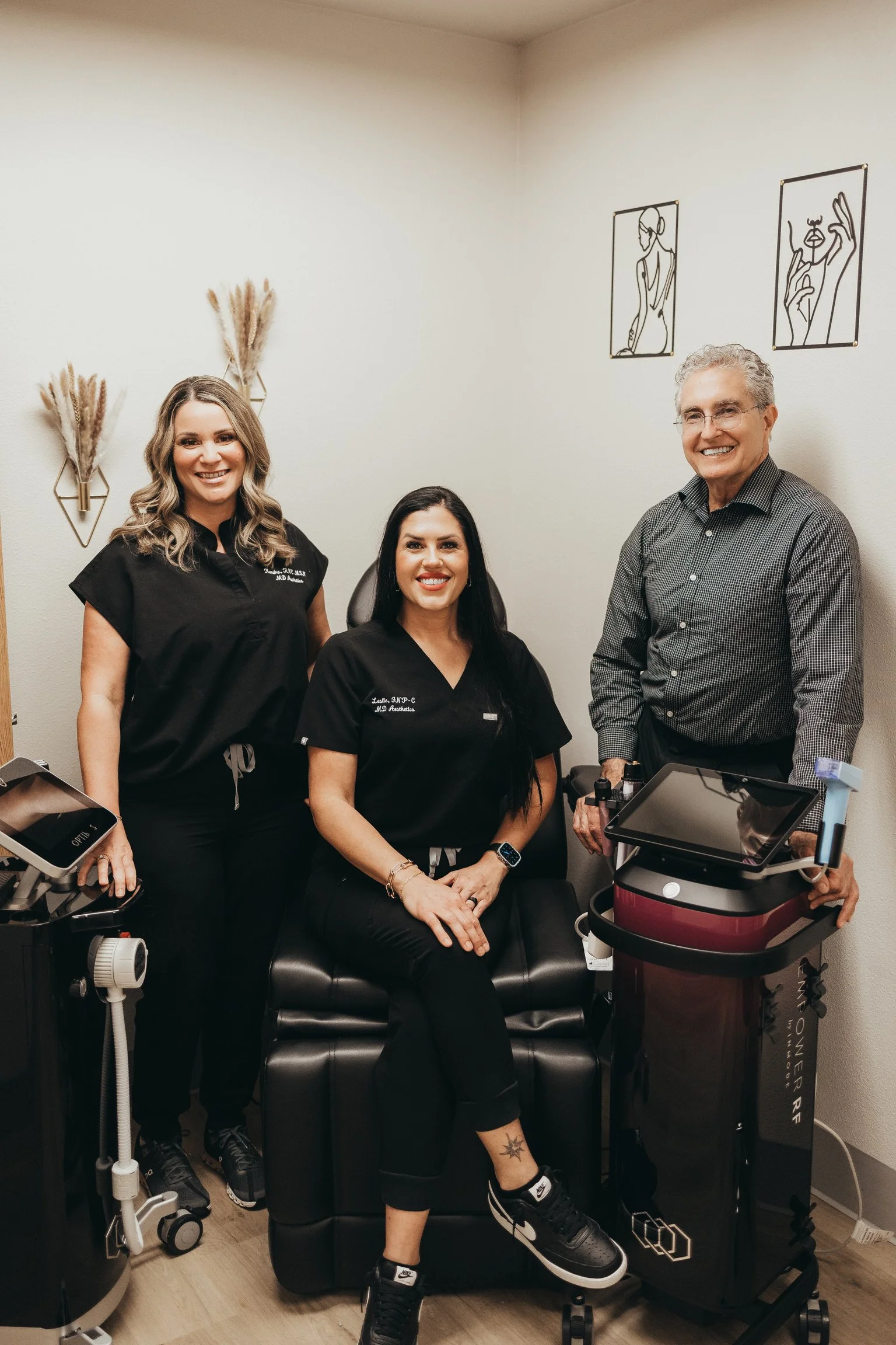 Three people in a medical or cosmetic office. Two women and one man, all smiling. The women are wearing black scrubs, and the man is in a gray checkered shirt. Medical equipment and modern wall art are visible.