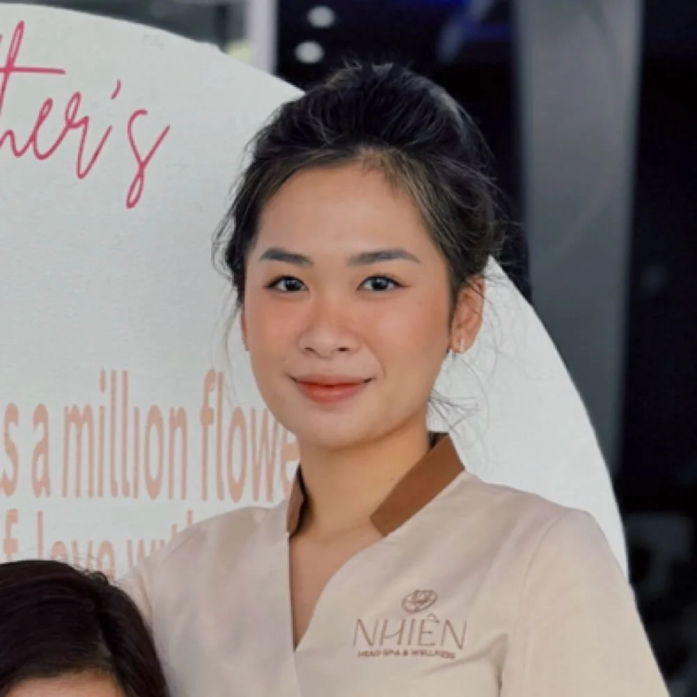 A young woman with dark hair tied back, wearing a beige uniform with a brown collar and the NHIEN logo, standing indoors in front of a white oval sign with orange and pink text.