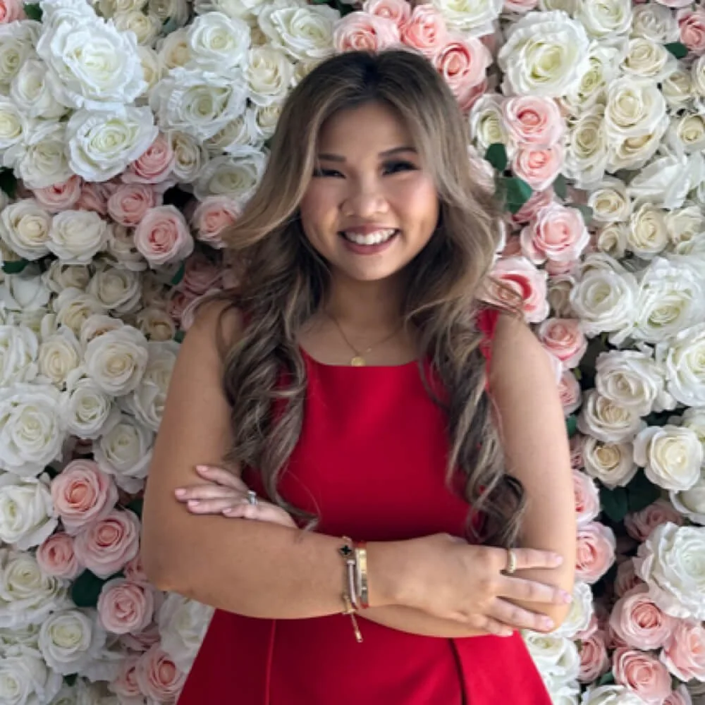 A woman with long wavy hair in a red dress standing in front of a wall of white and light pink roses, smiling with her arms crossed.