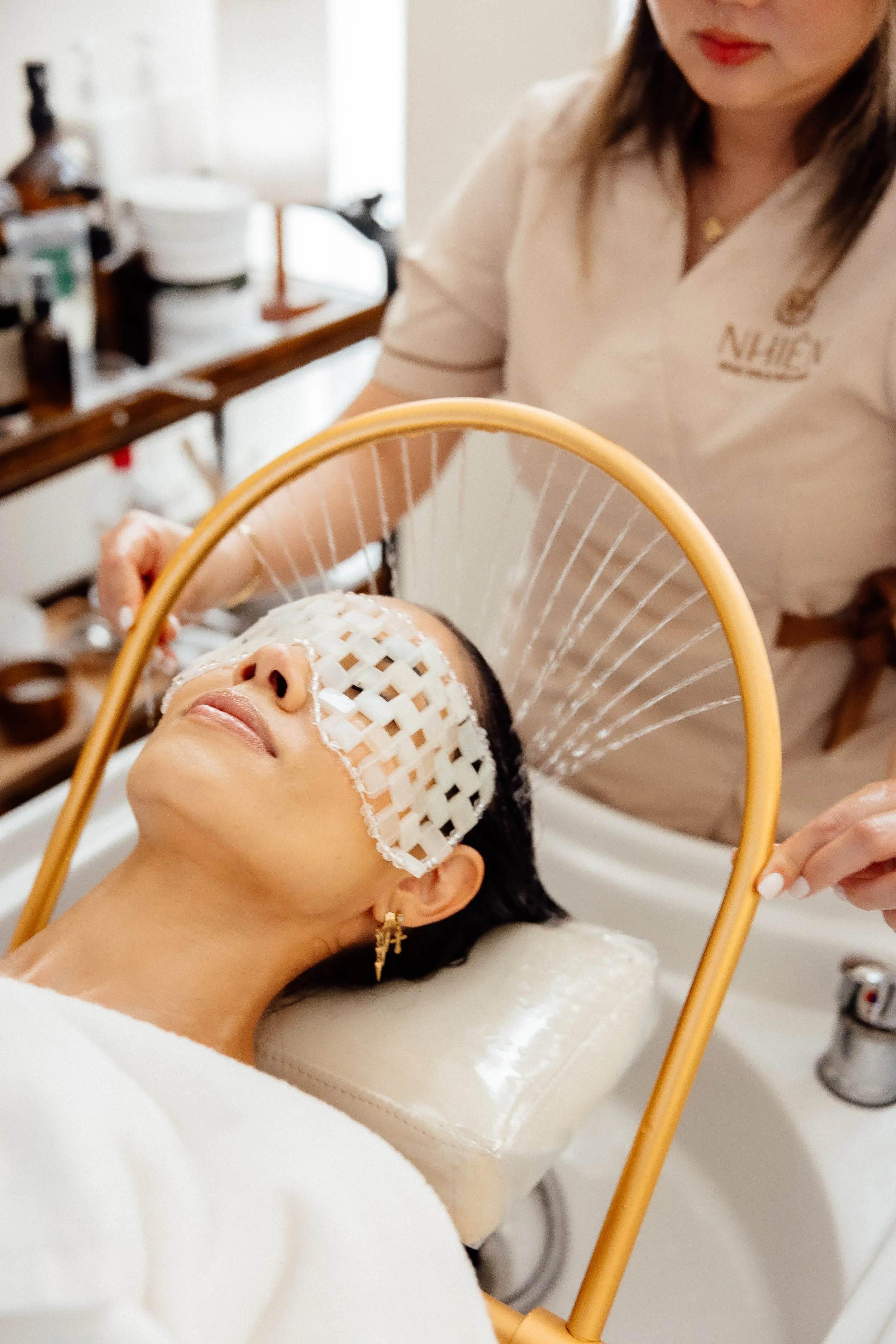 A woman receiving a facial treatment at a spa, with a spa therapist washing her hair in a hair washing sink.