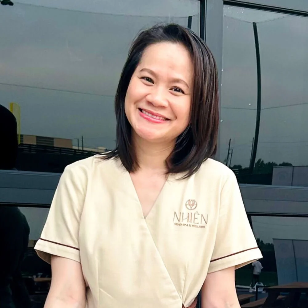 A woman with shoulder-length dark hair smiling outdoors, wearing a beige uniform with the logo and text 'NHIEN'.