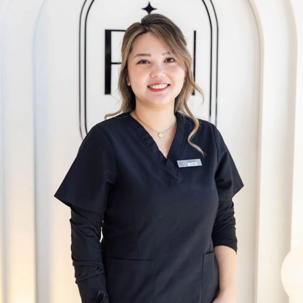 A woman wearing a black medical uniform standing in front of a decorative wall with the letter F. She is smiling and wearing a name tag.