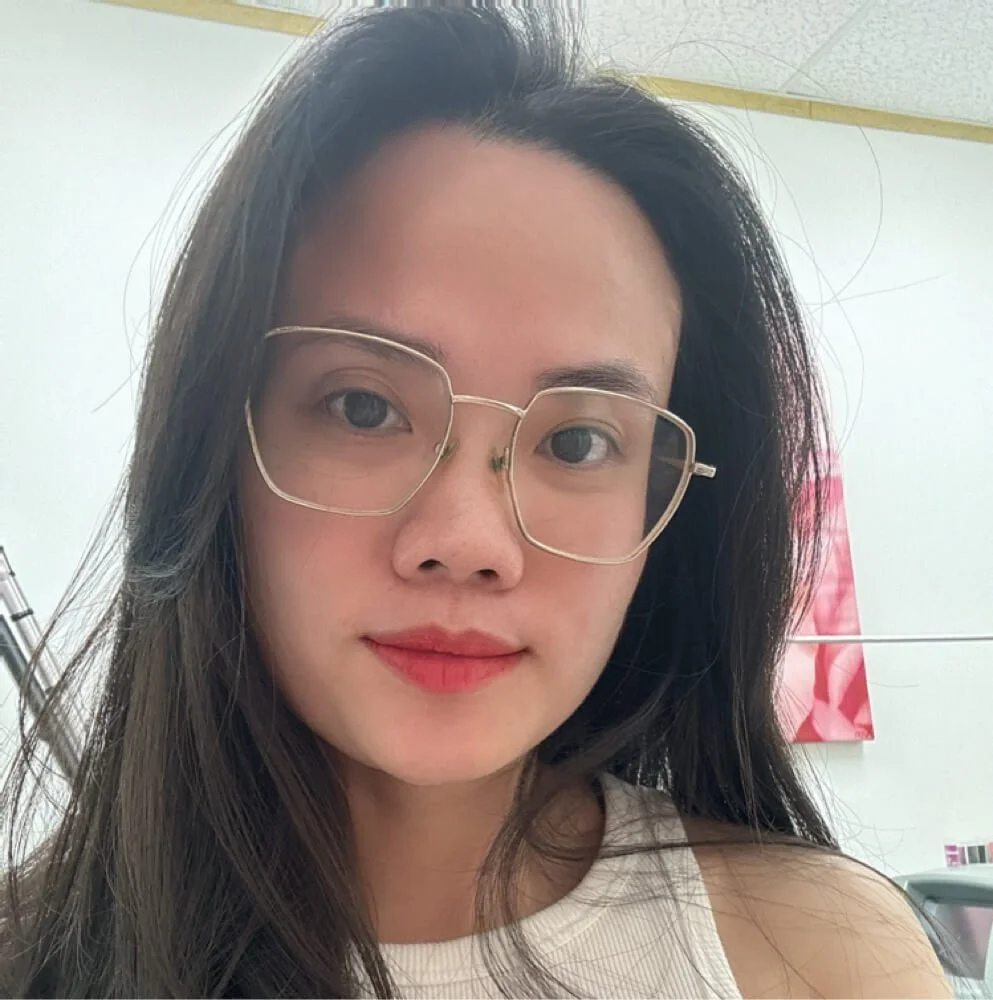 A young woman with long dark hair and glasses taking a selfie in a room with white walls and a pink painting in the background.