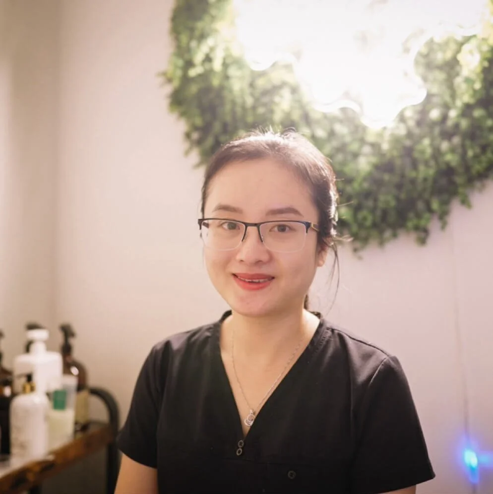 A woman with glasses and a black shirt smiling indoors with a leafy green wall decor and sunlight behind her.