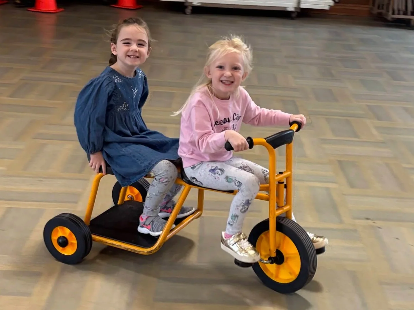 Two young girls are riding a yellow tricycle indoors on a wooden floor, smiling and enjoying their time.