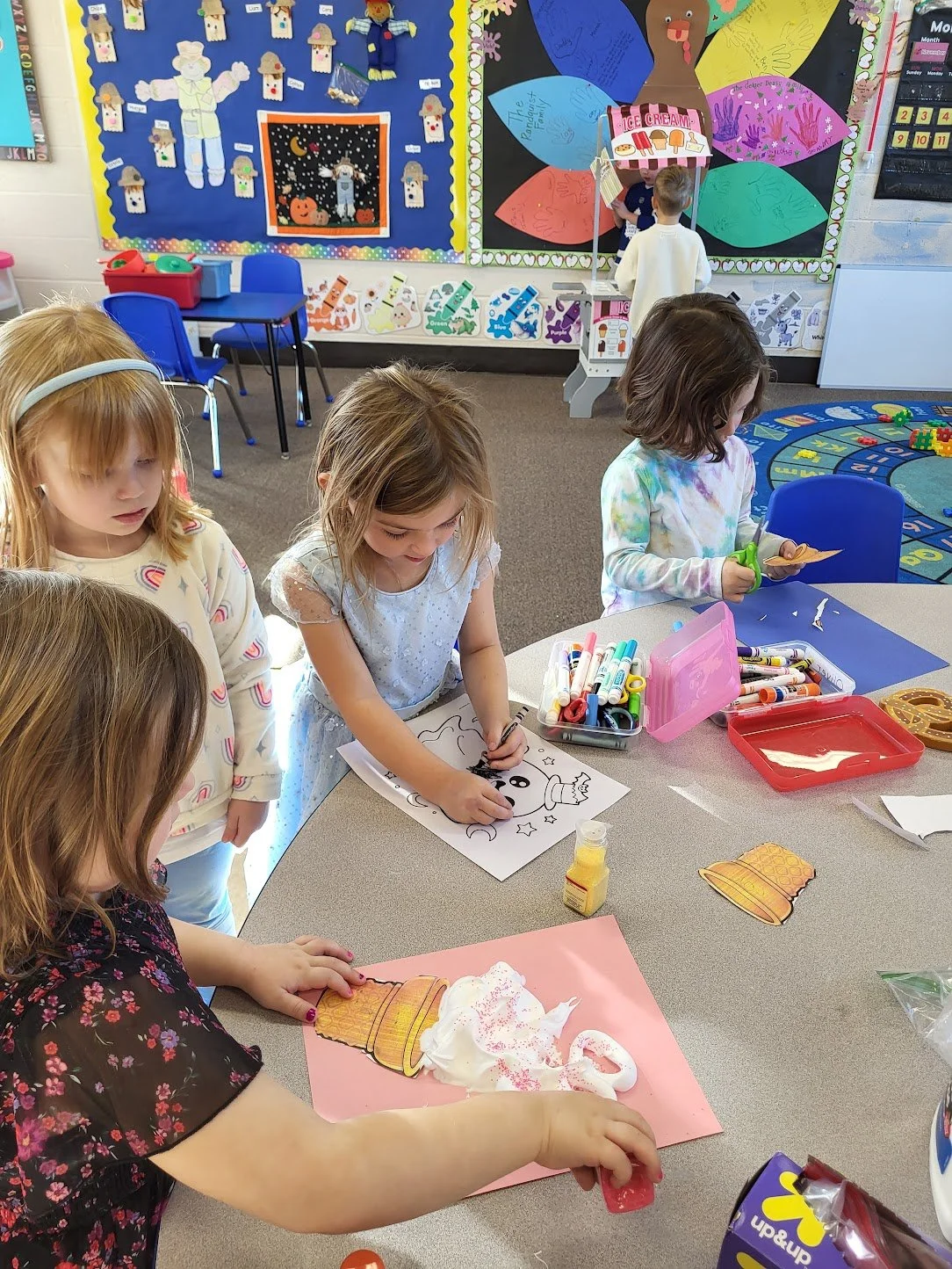 A group of young children engaged in arts and crafts at a classroom table with coloring pages, markers, and craft supplies, in a colorful classroom decorated with educational posters and a large display on the wall.