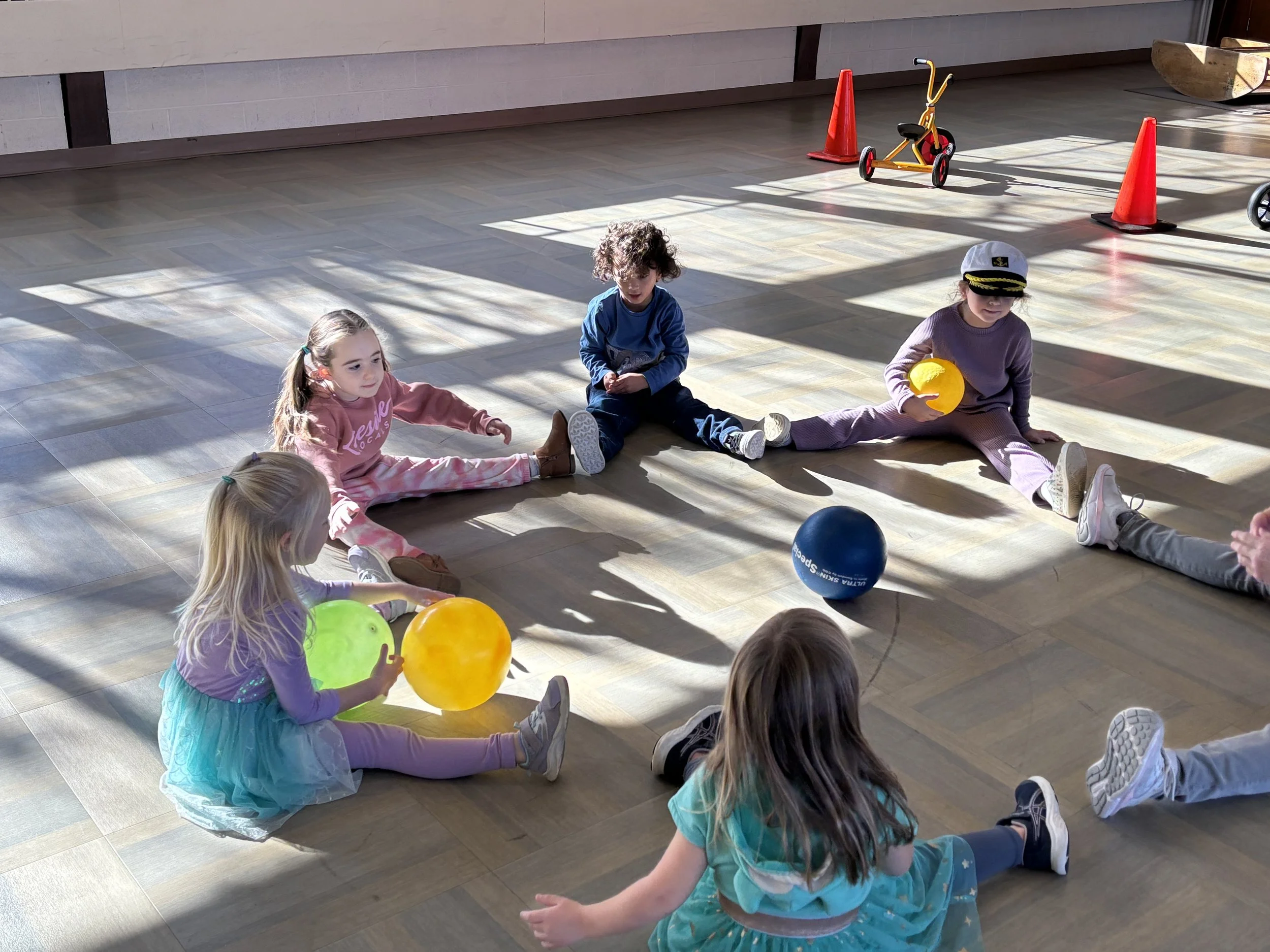 Children sitting on the floor in a circle in a play area, with some holding balloons and a ball, under sunlight with shadows cast on the floor.