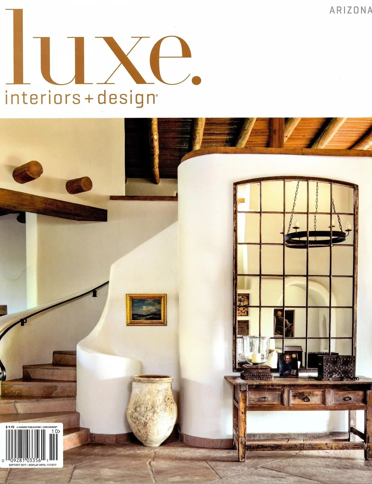 Luxe Interiors + Design magazine cover featuring an interior with a curved staircase, rustic wooden beams, large pottery vase, wooden table with decor, and a grid-patterned mirror reflecting a chandelier.