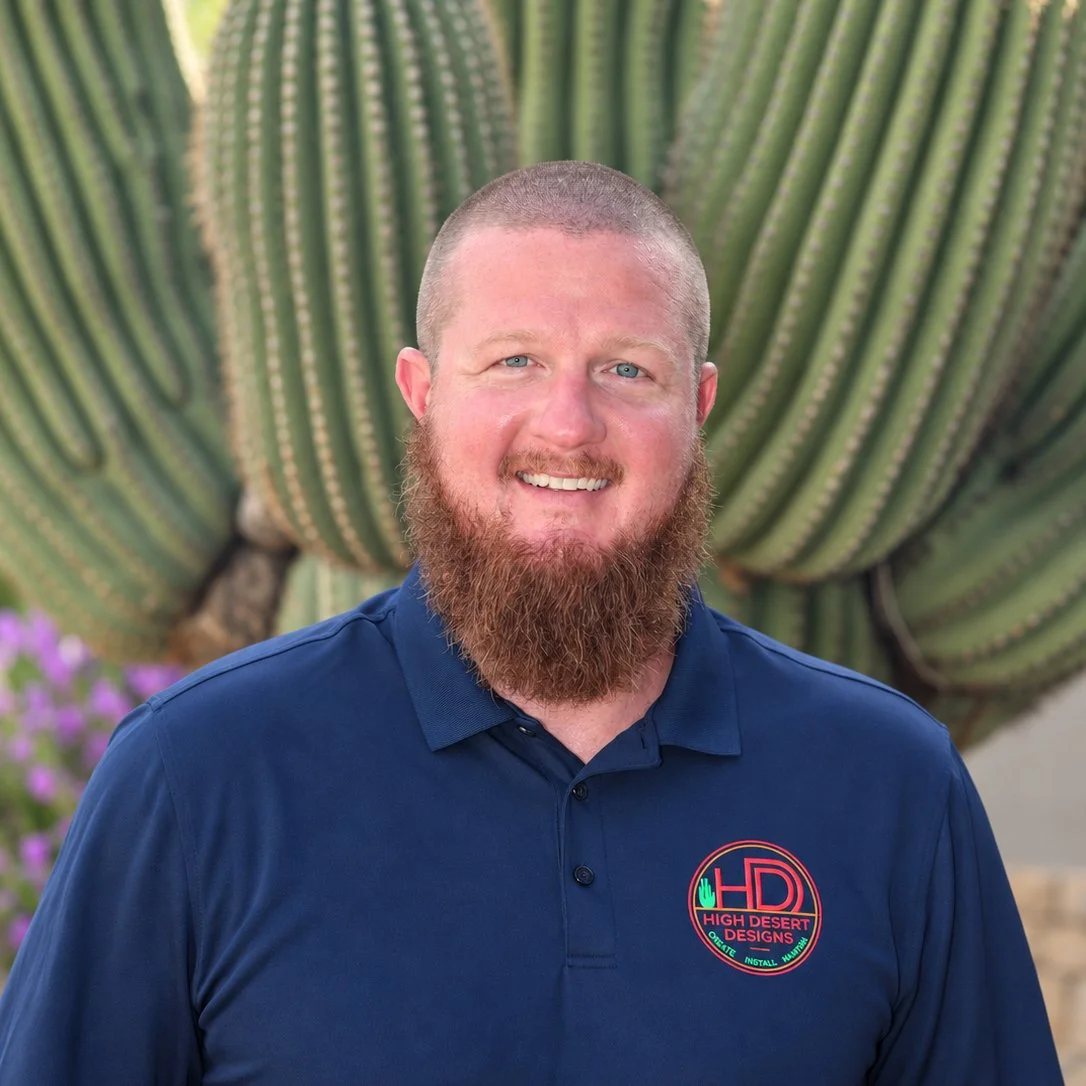 Man with curly hair smiling wearing a dark polo shirt with a logo, standing outdoors with blurred background.