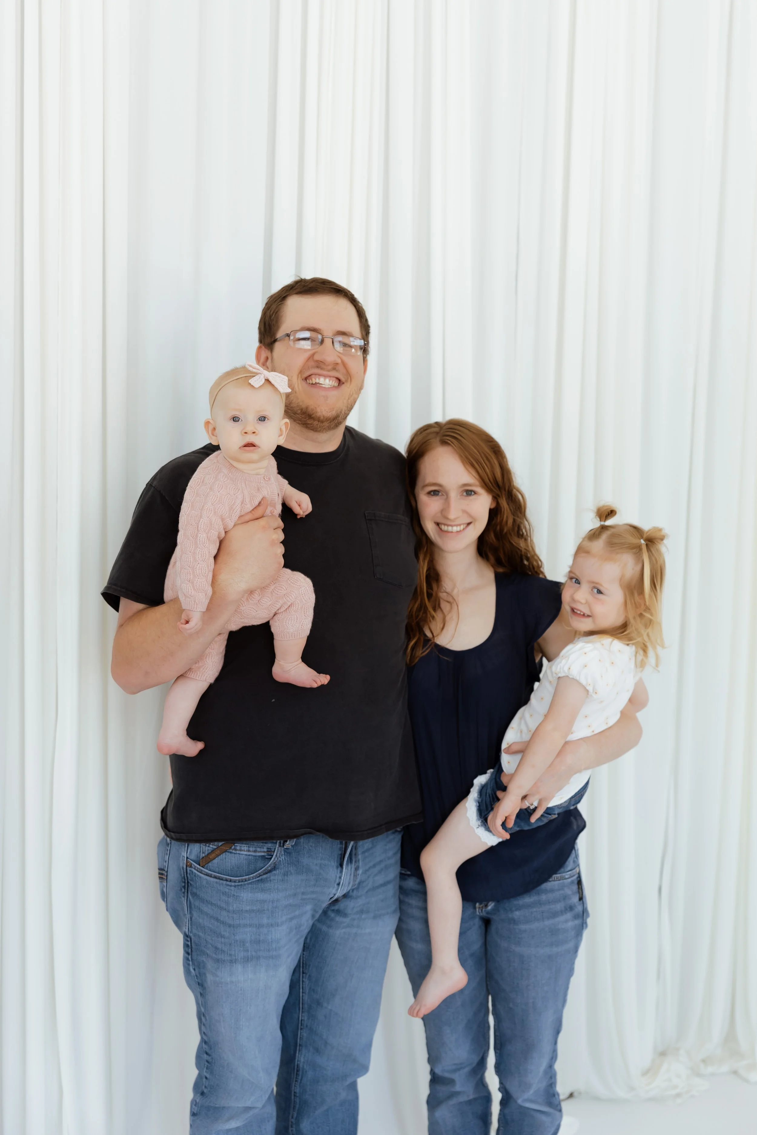 A smiling man holding a baby girl wearing a pink outfit, a woman with red hair holding a blonde girl, all standing in front of white curtains.