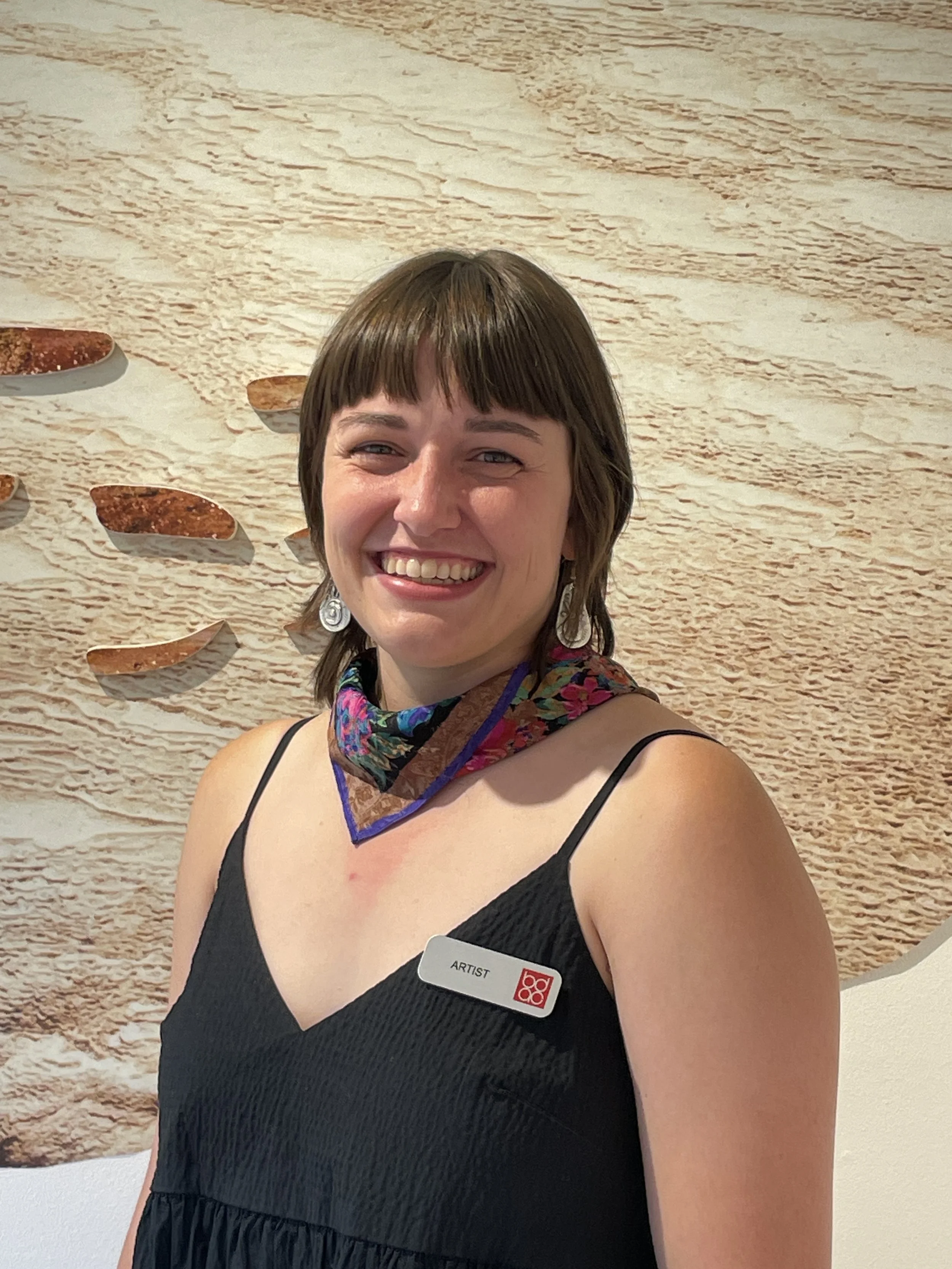 A woman smiling, wearing a black dress and a colorful scarf around her neck, standing in front of a textured beige wall with abstract art. She has shoulder-length brown hair and is wearing earrings. She has a name tag that reads "ARTIST".