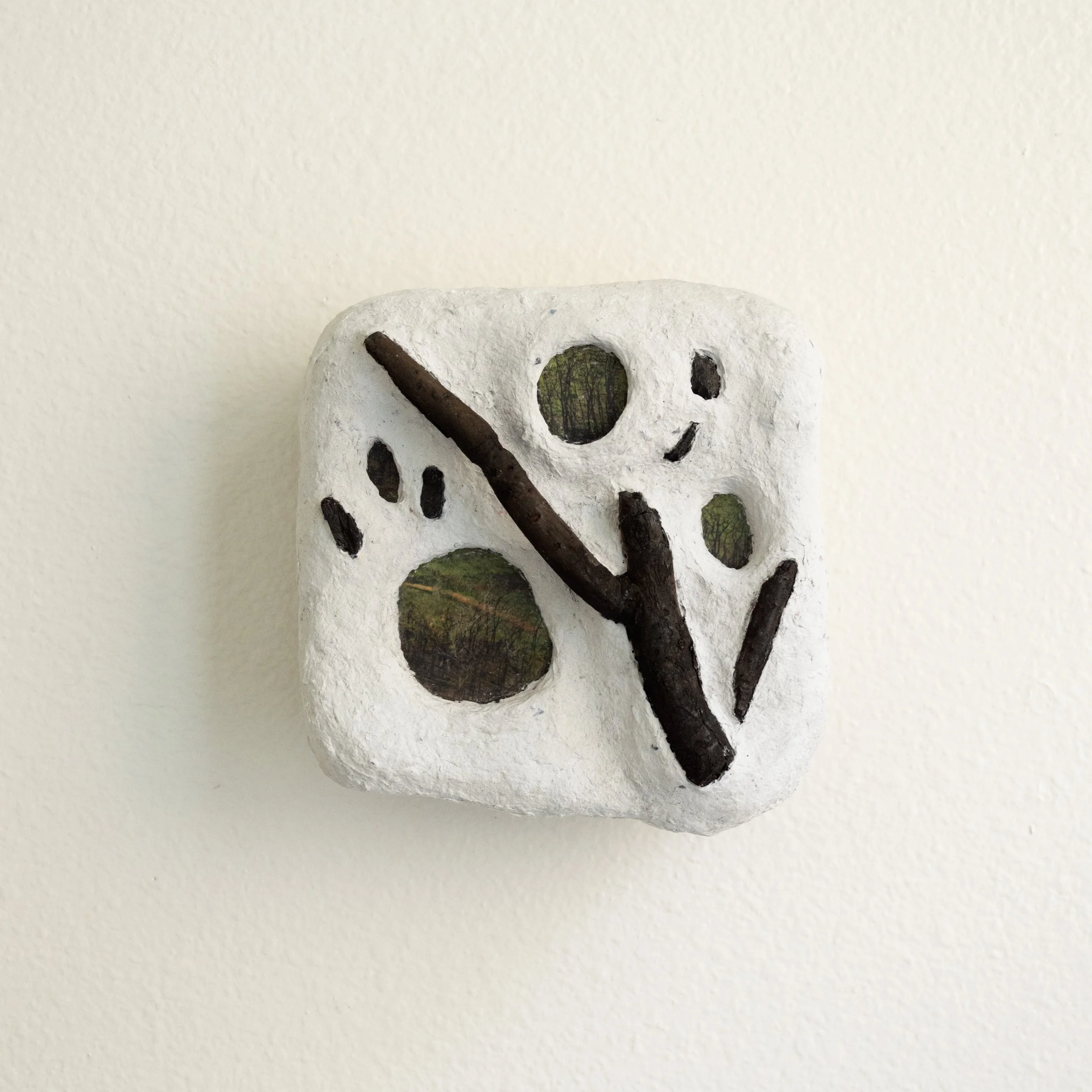 A white decorative object with holes, black branches, and green foliage inside some holes, set against a plain light background.