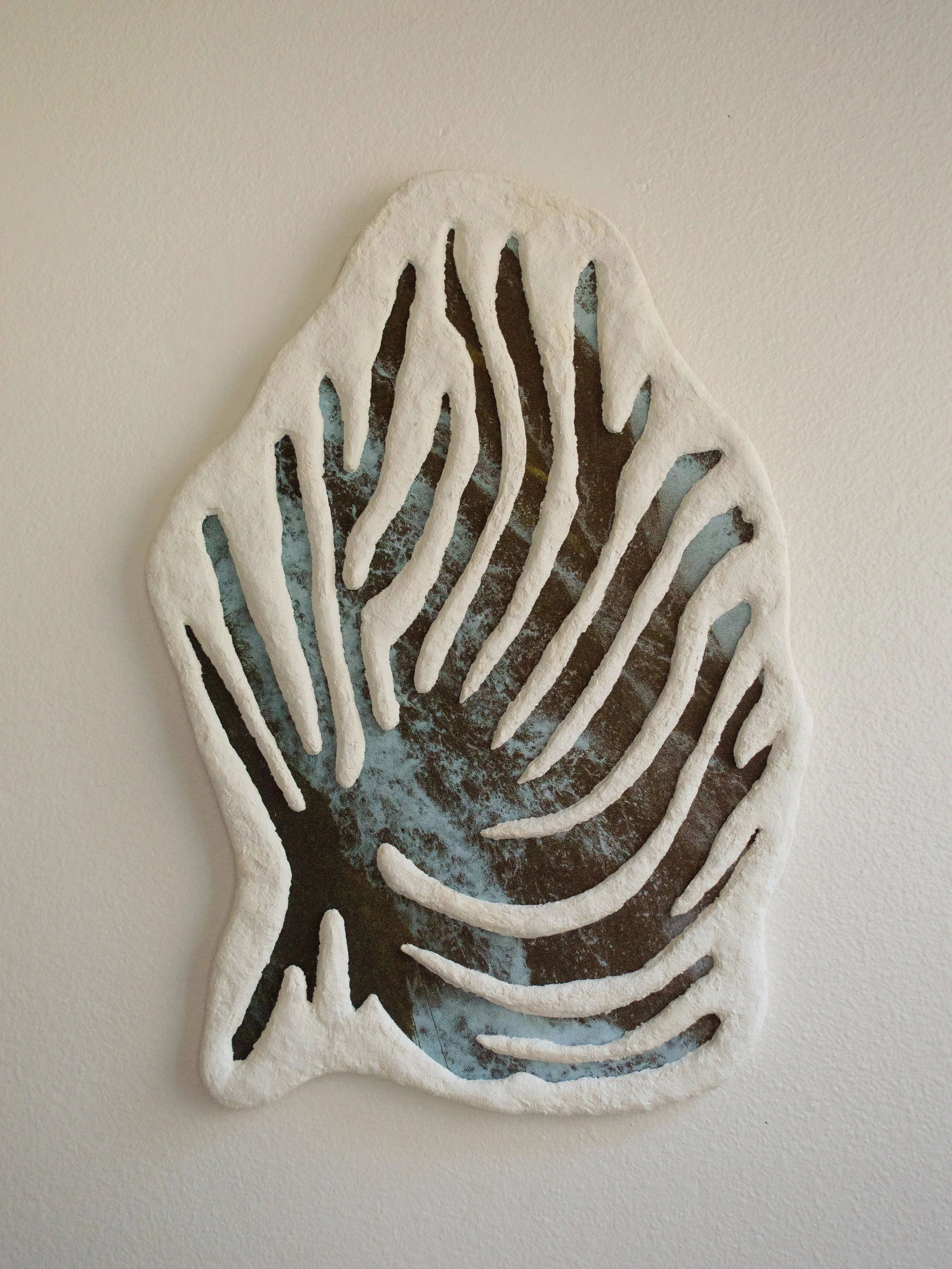 A decorative wall art piece of a leaf with cut-out veins, painted in white with some dark and blue accents visible through the cut-outs, mounted on a plain wall.