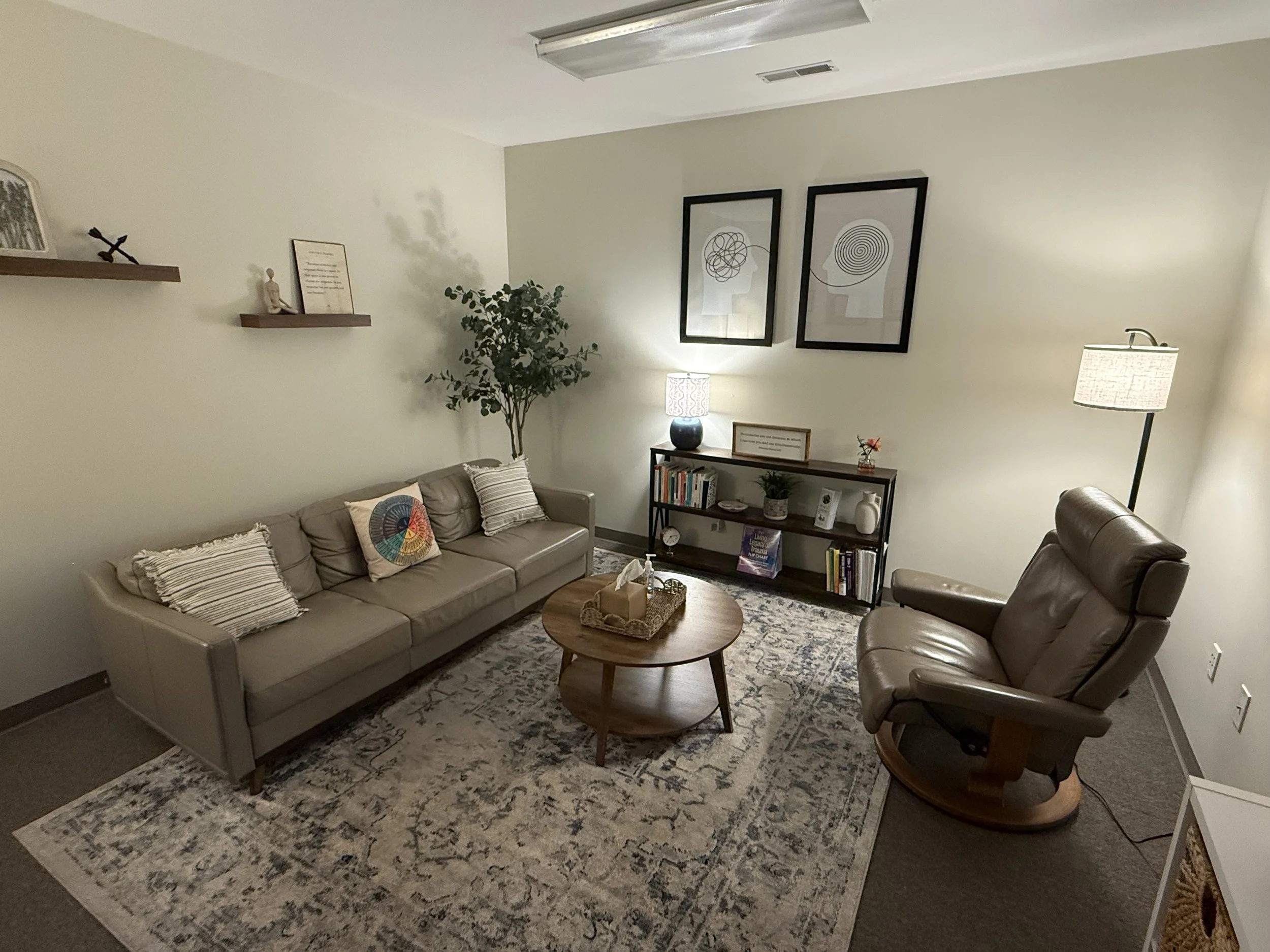 Living room with beige sofa, navy armchair, wooden coffee table, patterned rug, bookshelf with plants and books, wall art, and decorative shelves with picture frames and small sculptures.