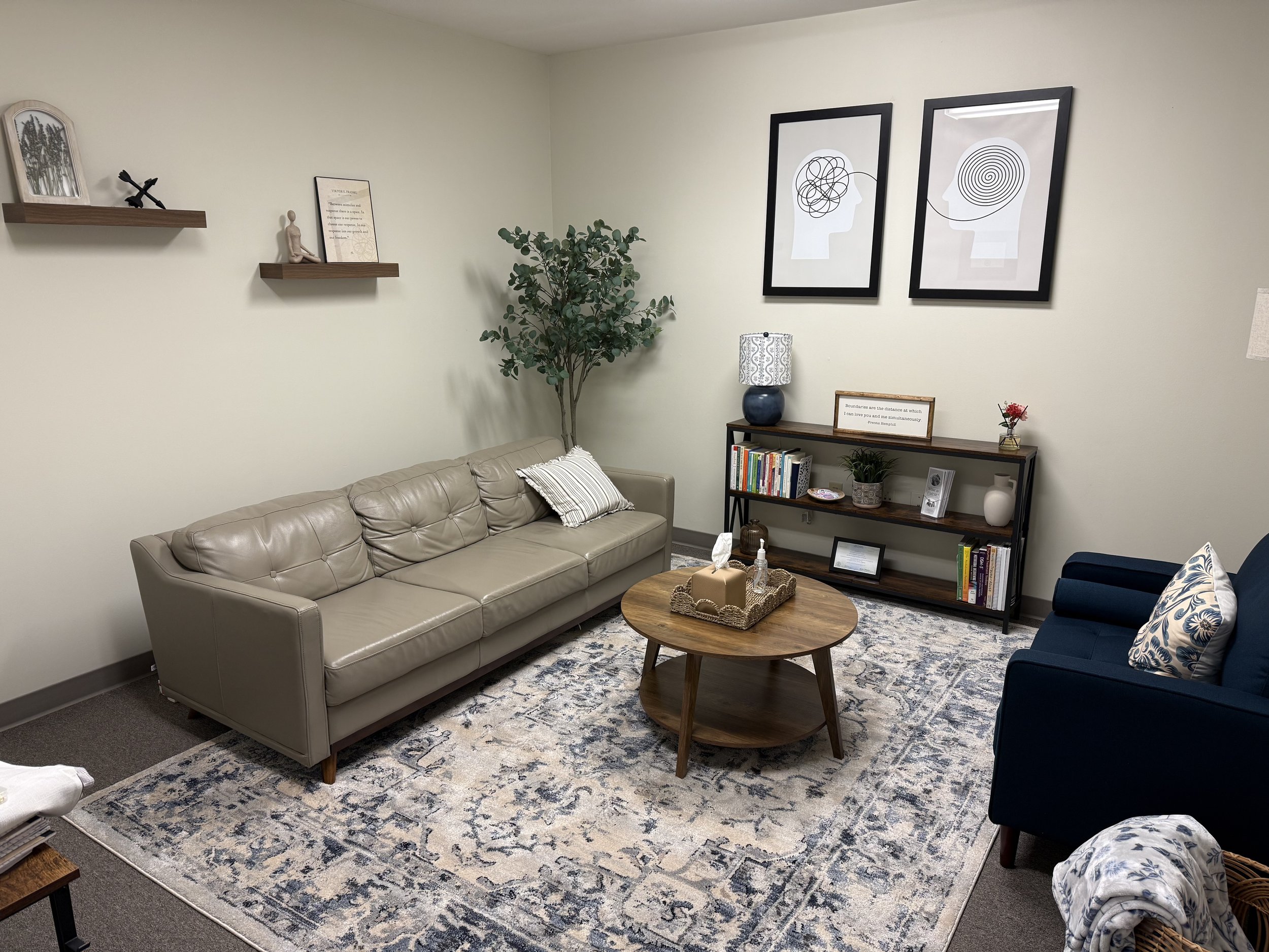 Living room with beige sofa, navy armchair, wooden coffee table, patterned rug, bookshelf with plants and books, wall art, and decorative shelves with picture frames and small sculptures.