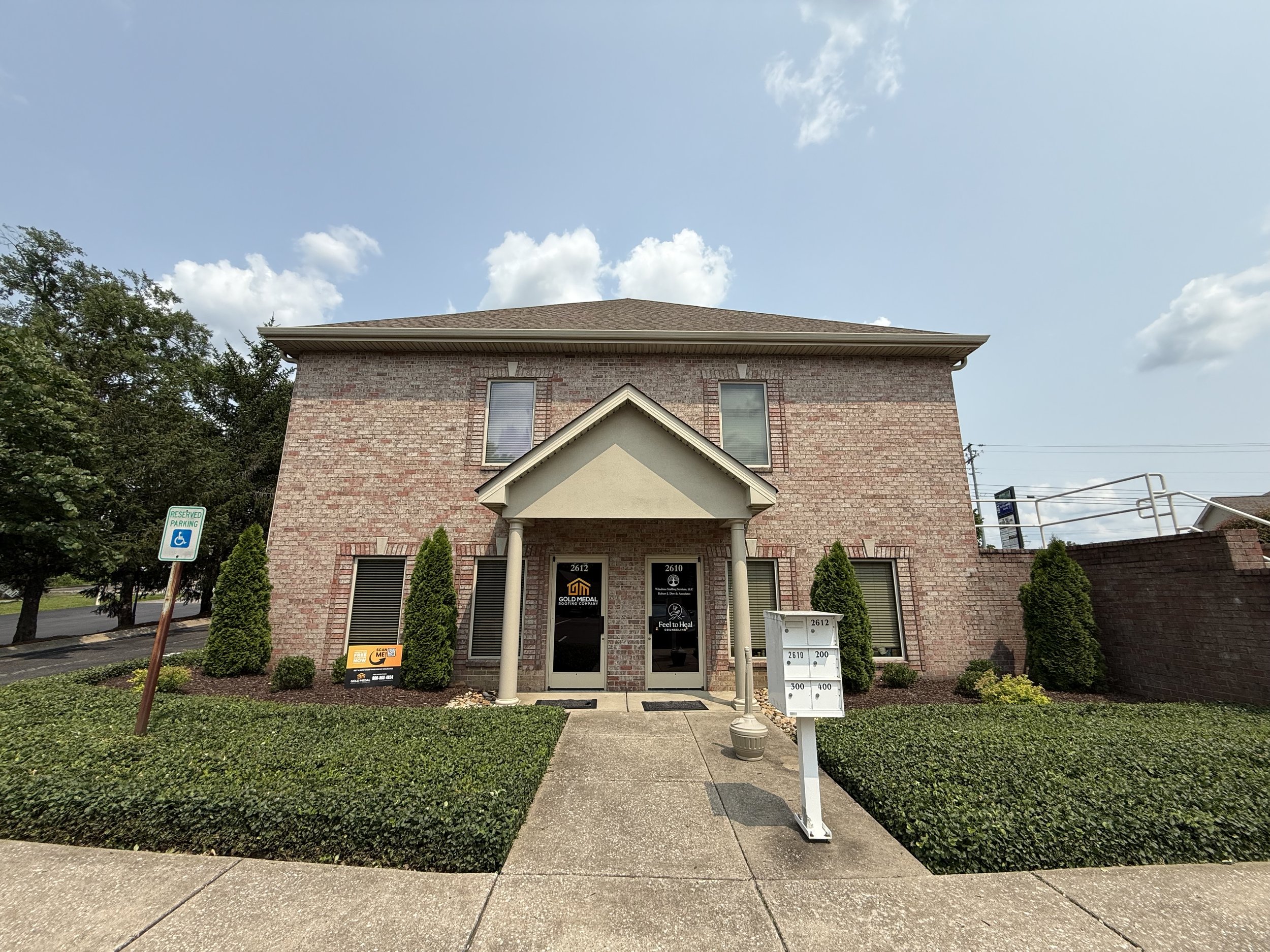 A brick building with a small porch, surrounded by neatly trimmed bushes, with business signs on the door and sidewalk, and a reserved parking sign for handicapped