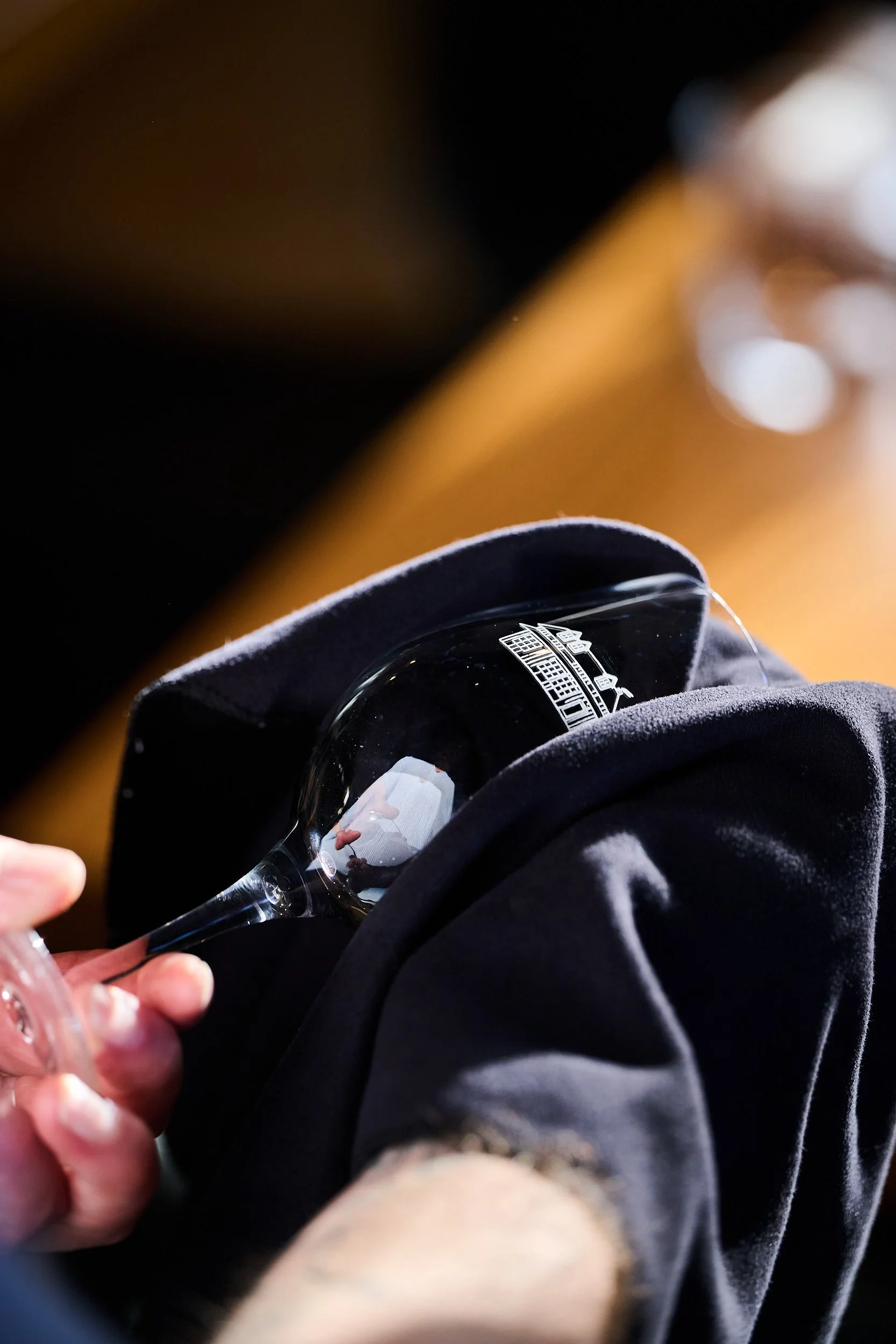 Personne tenant un verre à vin reflétant une personne prenant une photo, avec un motif brodé représentant un paysage urbain sur le verre, assise sur un manteau noir.