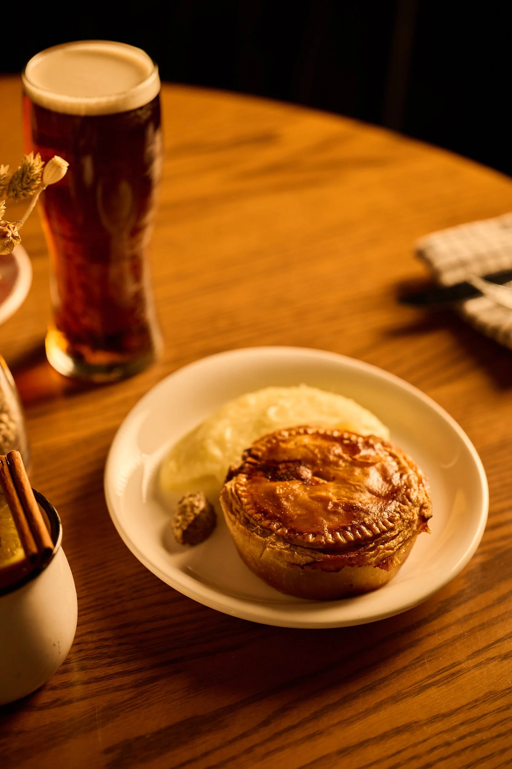 Une assiette avec une tourte à la viande, de la purée de pommes de terre et une petite portion de relish sur une table en bois, avec un verre de bière brune en arrière-plan.