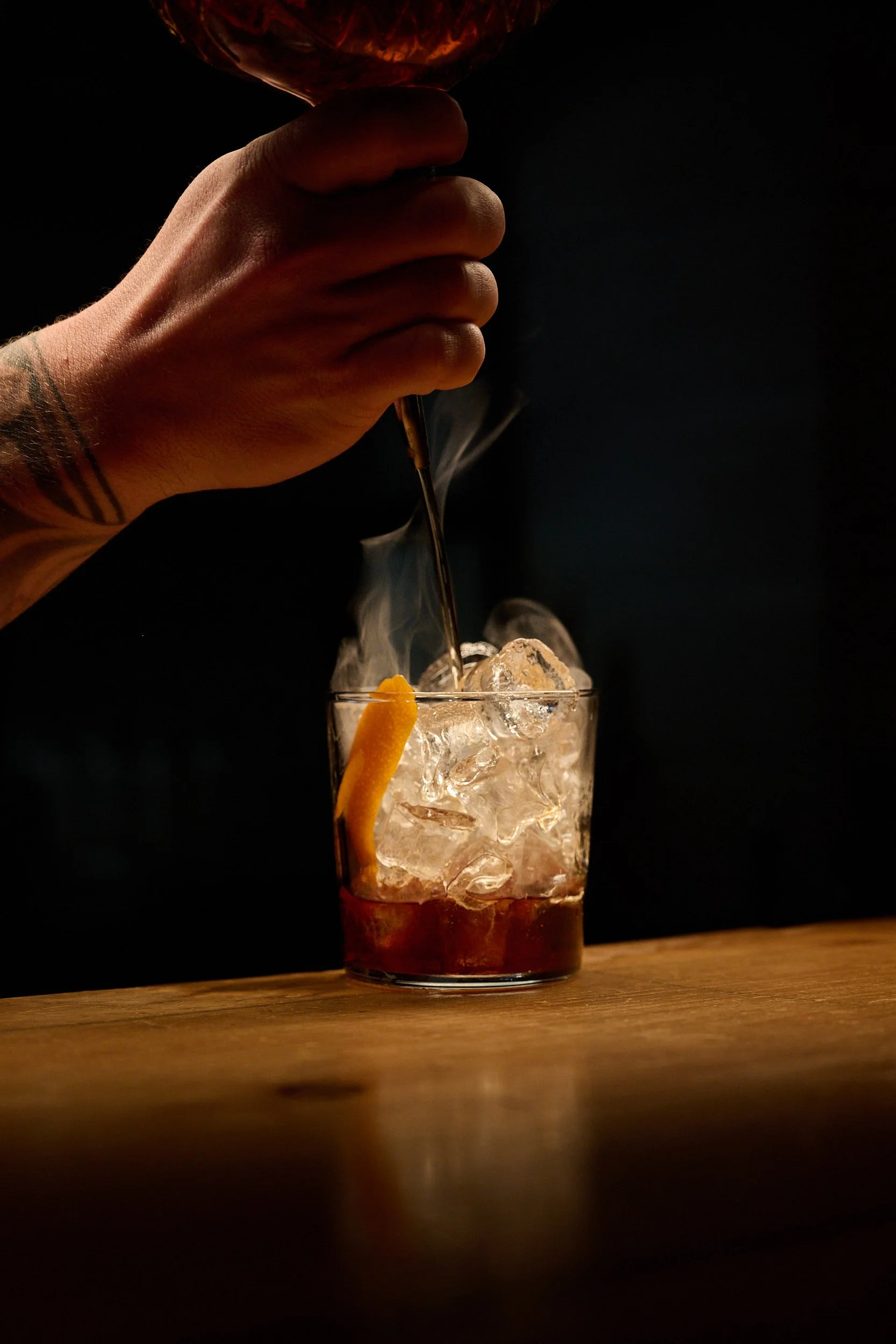 Une personne au bras tatoué verse une boisson dans un verre rempli de glaçons, garni d'un zeste de citron, sur un comptoir de bar en bois.
