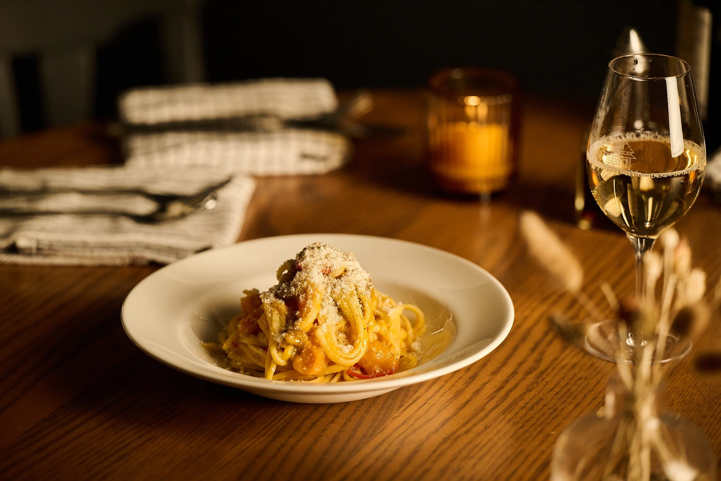 Une assiette de spaghettis recouverte de parmesan sur une table en bois, avec un verre de vin blanc et une bougie allumée en arrière-plan.