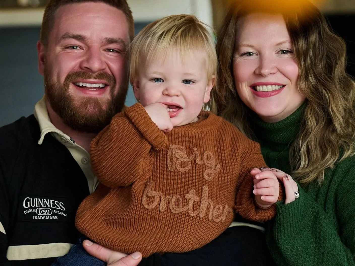 Tim Robertson and Lindsay Hughes holding a young child with blonde hair and blue eyes, in a cozy setting.