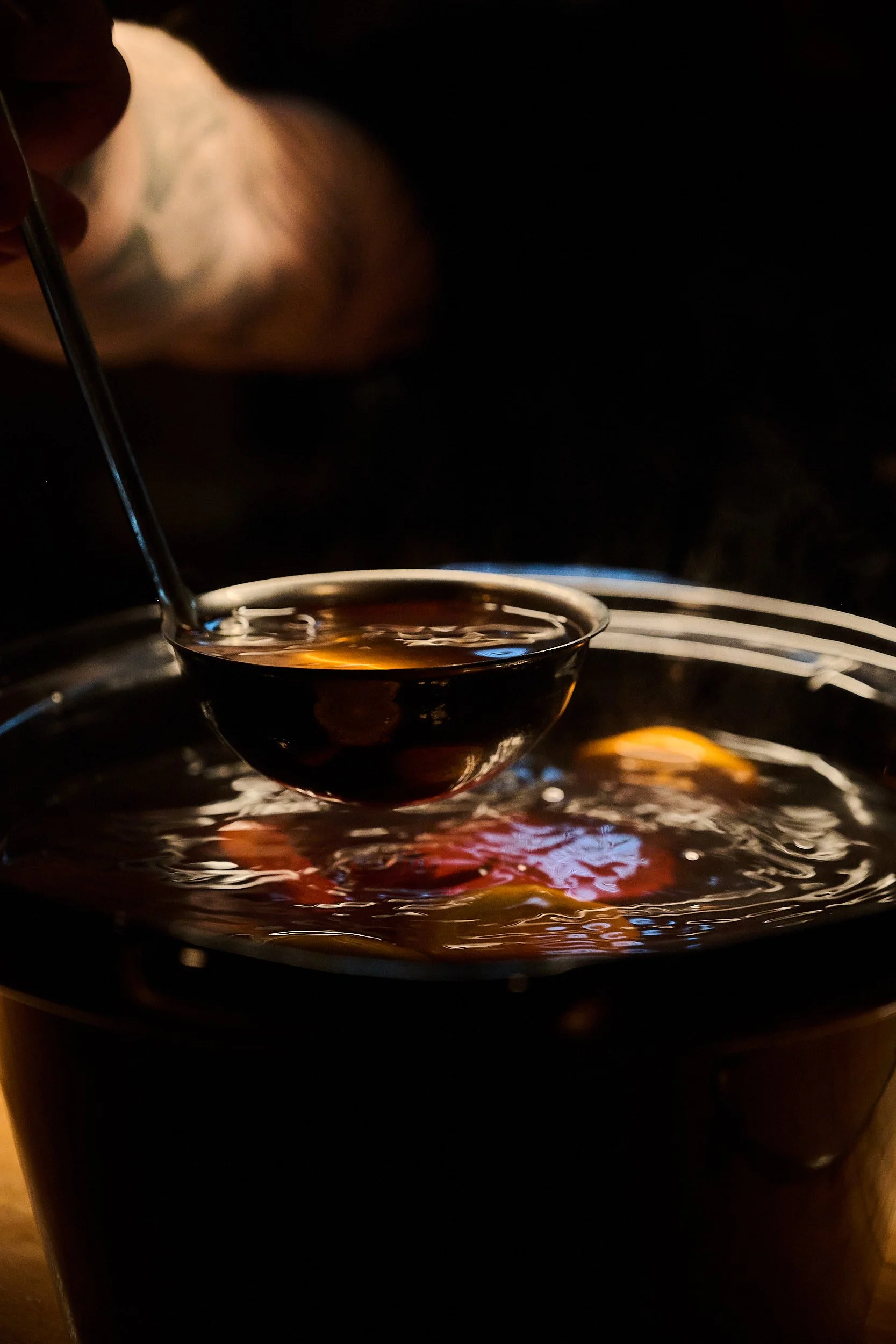 Une cuillère remplie d'un liquide sombre est prélevée dans un récipient. Le liquide reflète la lumière et laisse apparaître des objets colorés au fond du récipient.