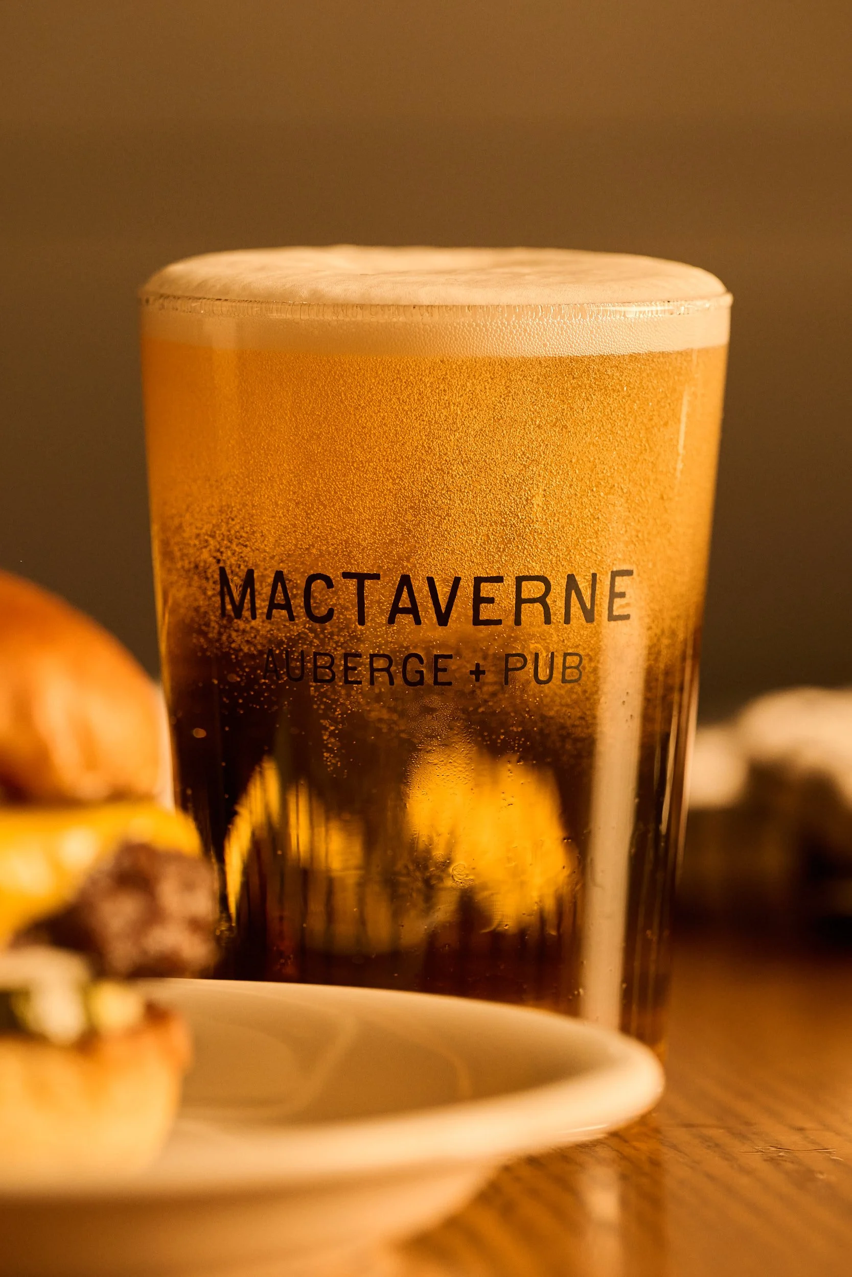 Un verre de bière avec de la mousse sur le dessus, portant l'inscription « MACTAVERNE AUBERGE + PUB », posé sur une table en bois avec un arrière-plan flou.