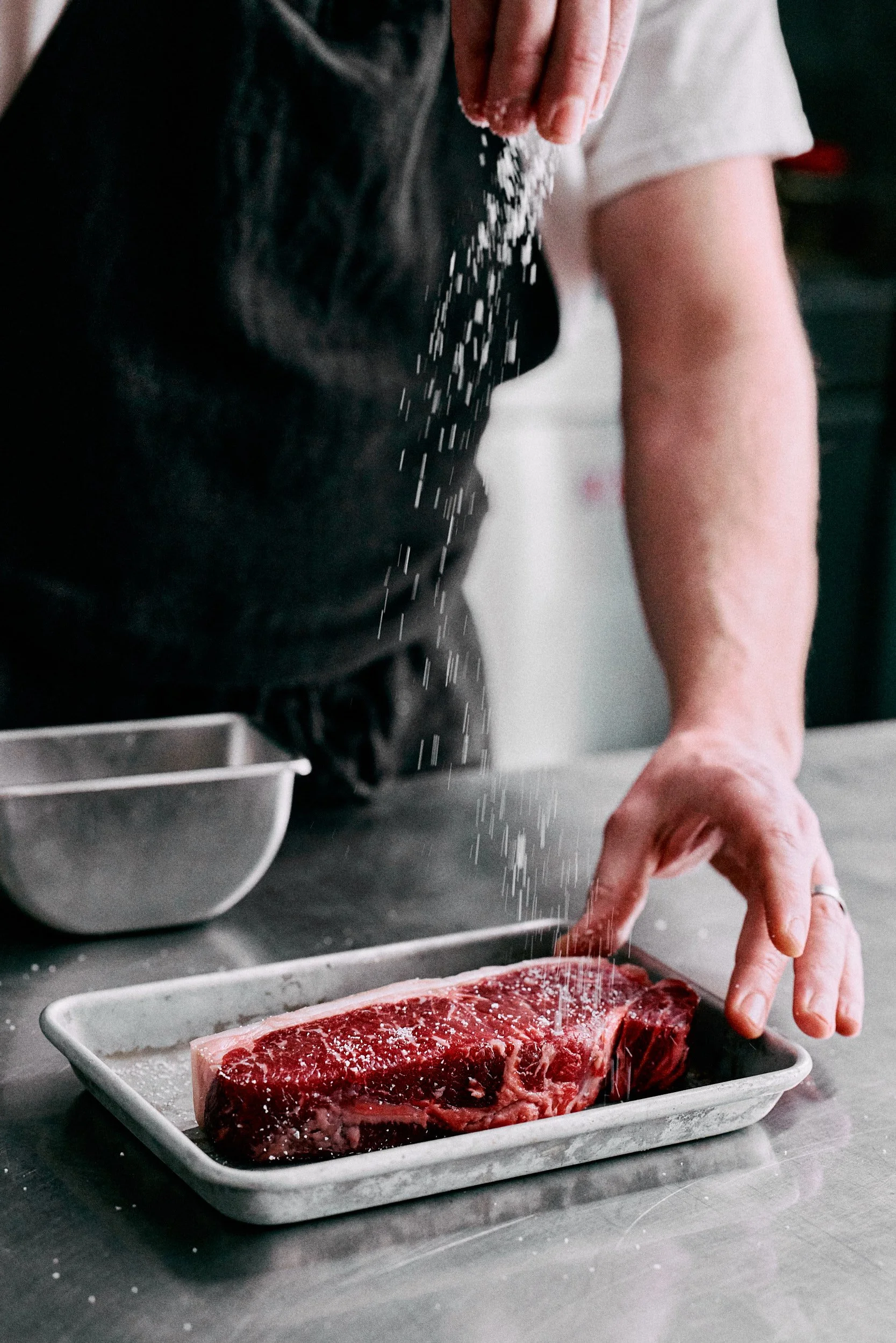 Une personne saupoudrant du sel sur un steak cru posé sur un plateau blanc dans une cuisine.