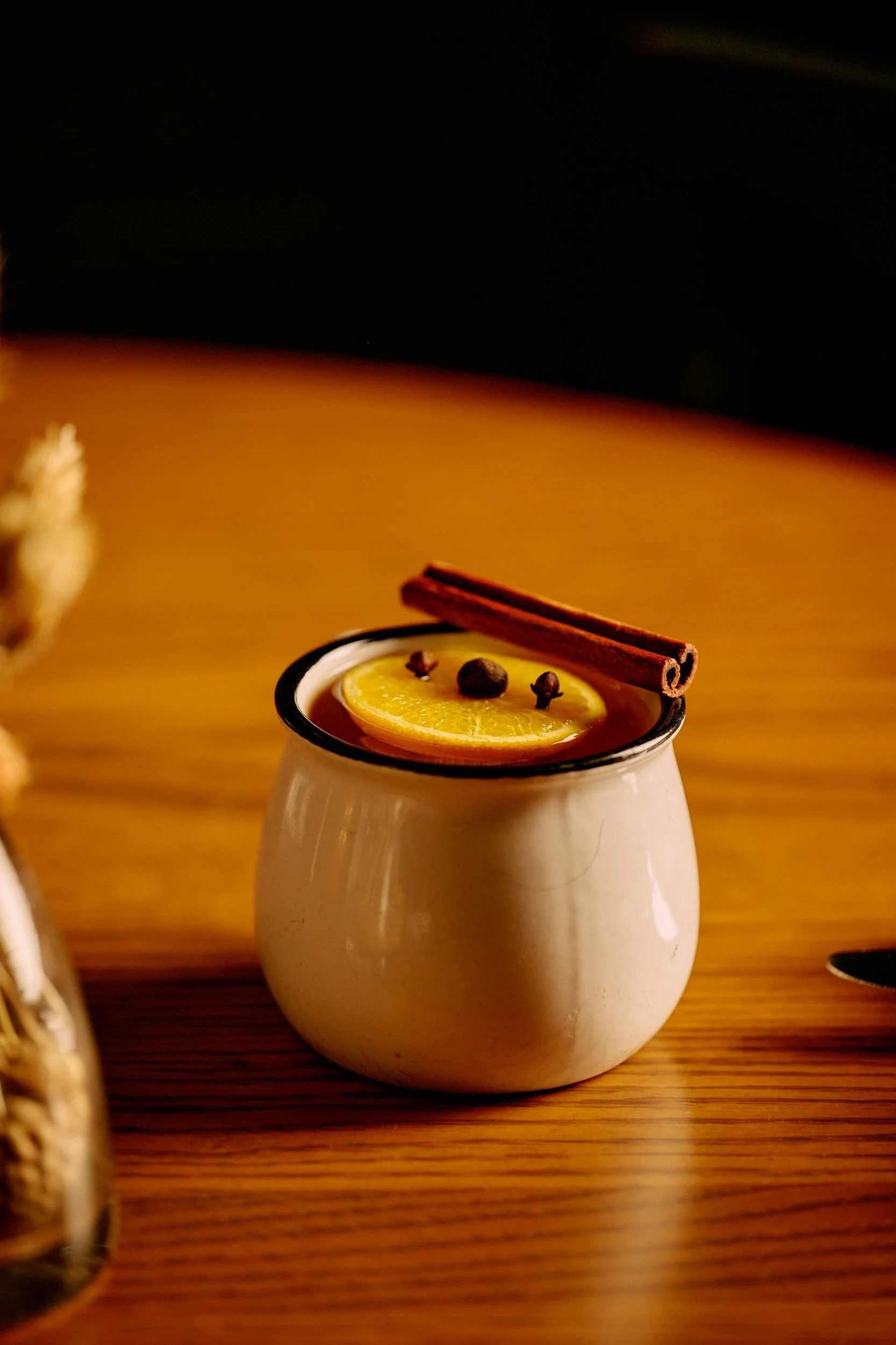 Une tasse en céramique blanche avec un rebord noir, remplie d'une boisson chaude garnie d'une tranche de citron, d'un bâton de cannelle et de grains de poivre entiers, posée sur une surface en bois.