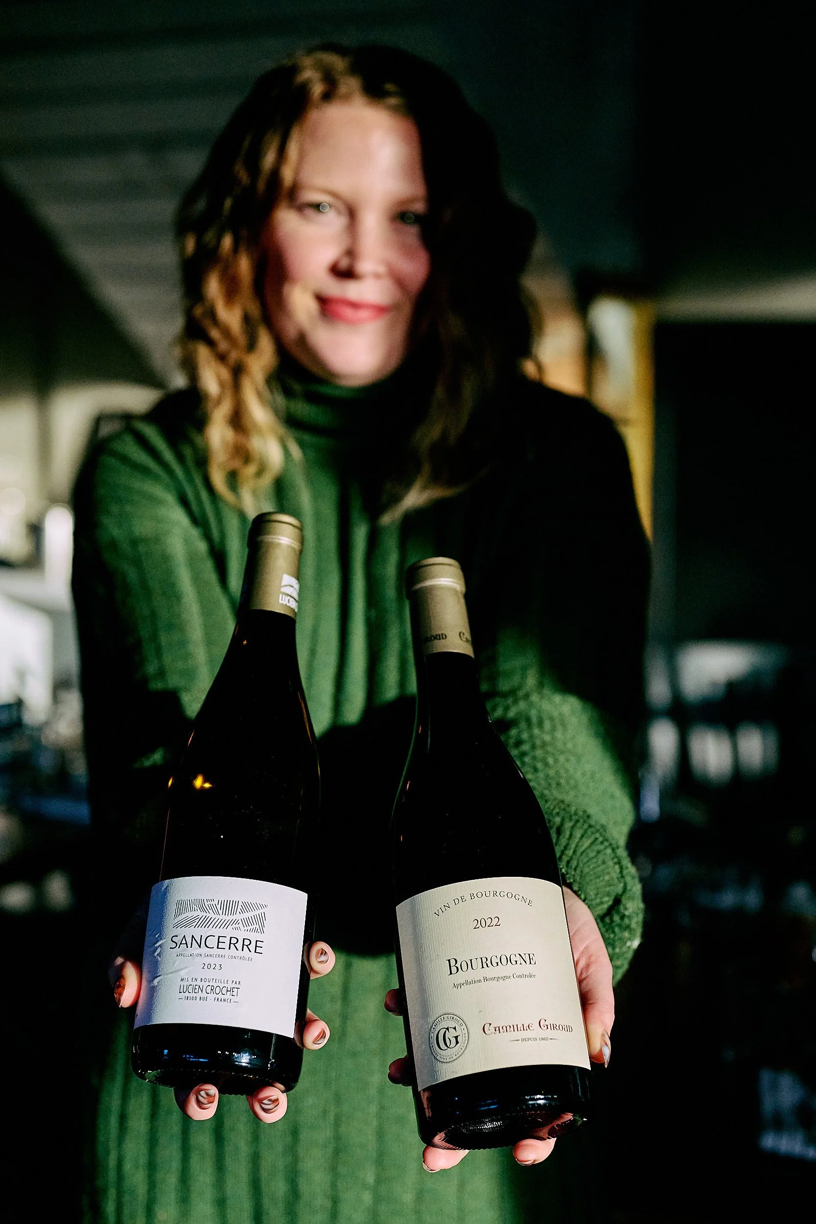 Femme aux cheveux bruns ondulés, vêtue d'un col roulé vert foncé, tenant deux bouteilles de vin, debout à l'intérieur avec un arrière-plan sombre et légèrement flou.