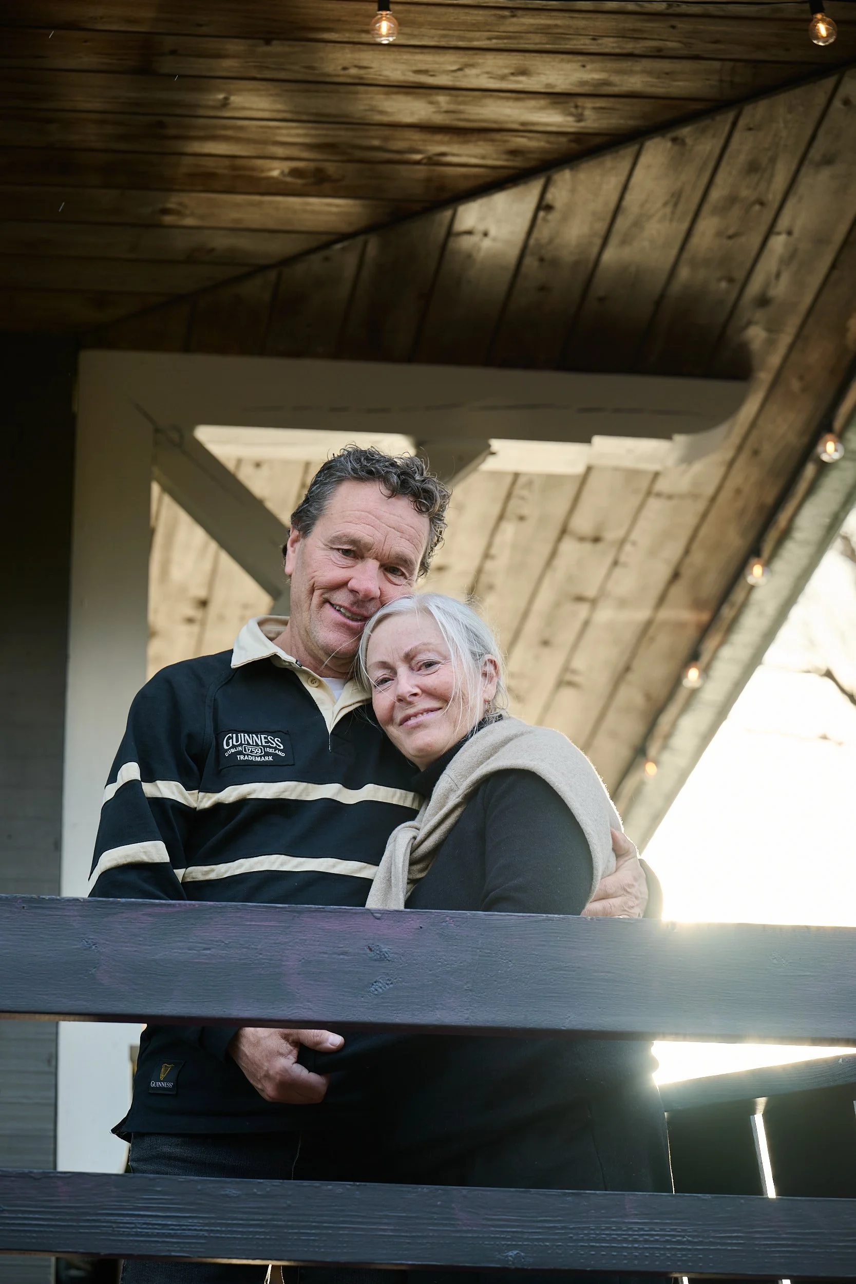 Craig Robertson and Lynne Robertson embrace outdoors near MacTaverne with a sloped roof, smiling warmly wearing a Guinness shirt in Val-david. 