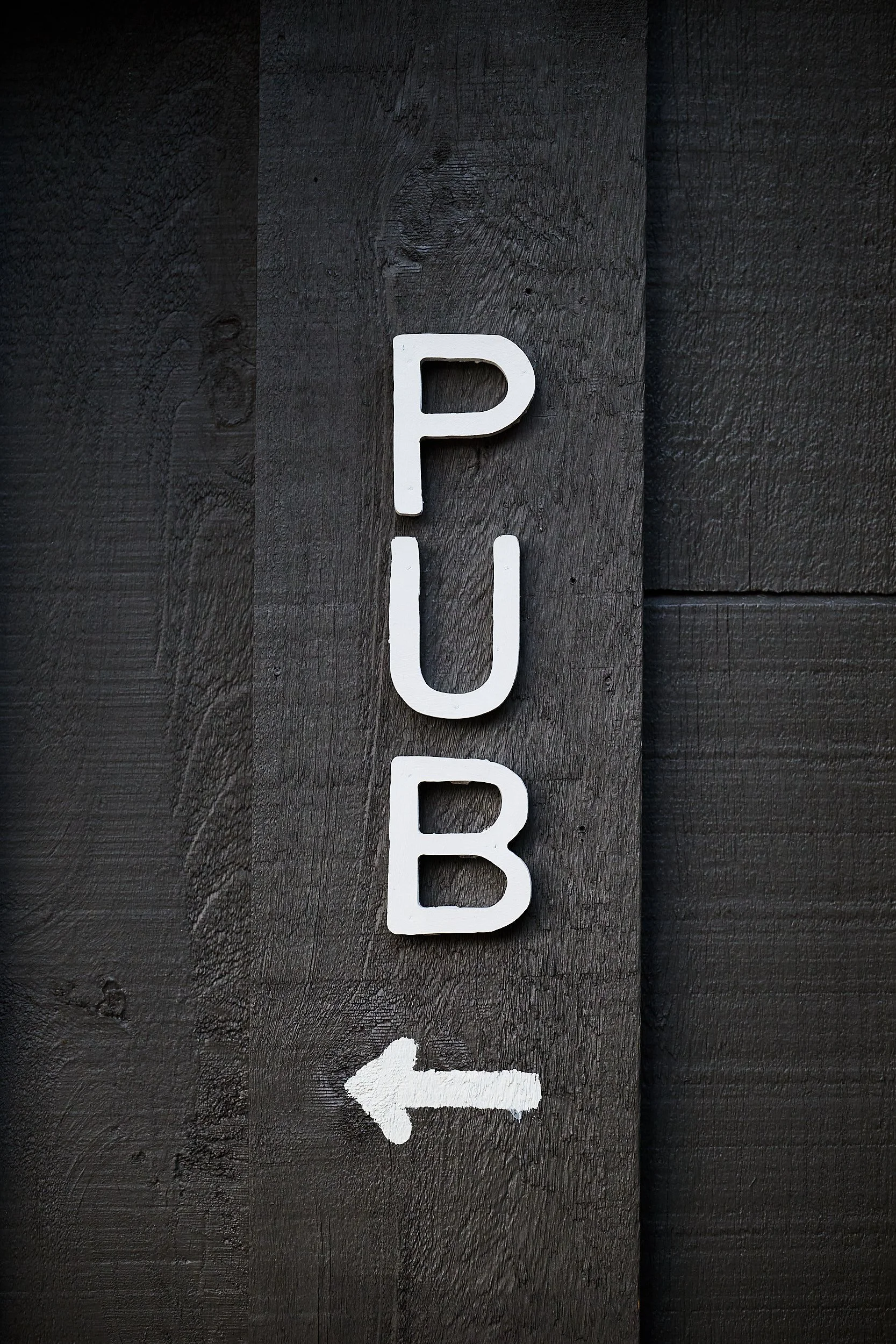 Panneau peint en blanc indiquant « PUB » avec une flèche pointant vers la gauche, fixé sur un mur en bois foncé.