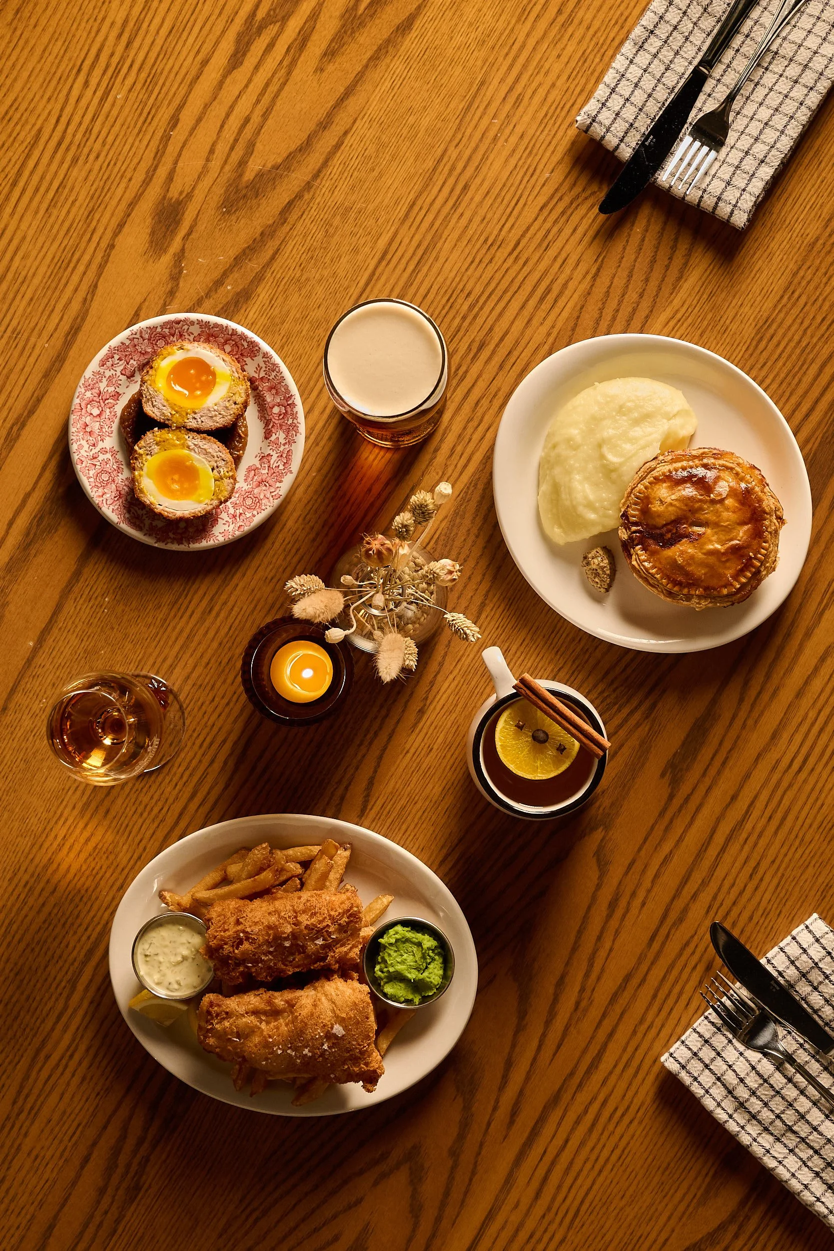 Vue plongeante sur une table à manger en bois dressée pour un repas, avec des assiettes contenant notamment de la purée de pommes de terre, une tarte salée, du poulet frit accompagné de frites et de condiments, ainsi que des verres de bière, une tasse de thé chaud au citron, de petites bougies, une serviette avec des couverts et un arrangement floral décoratif.
