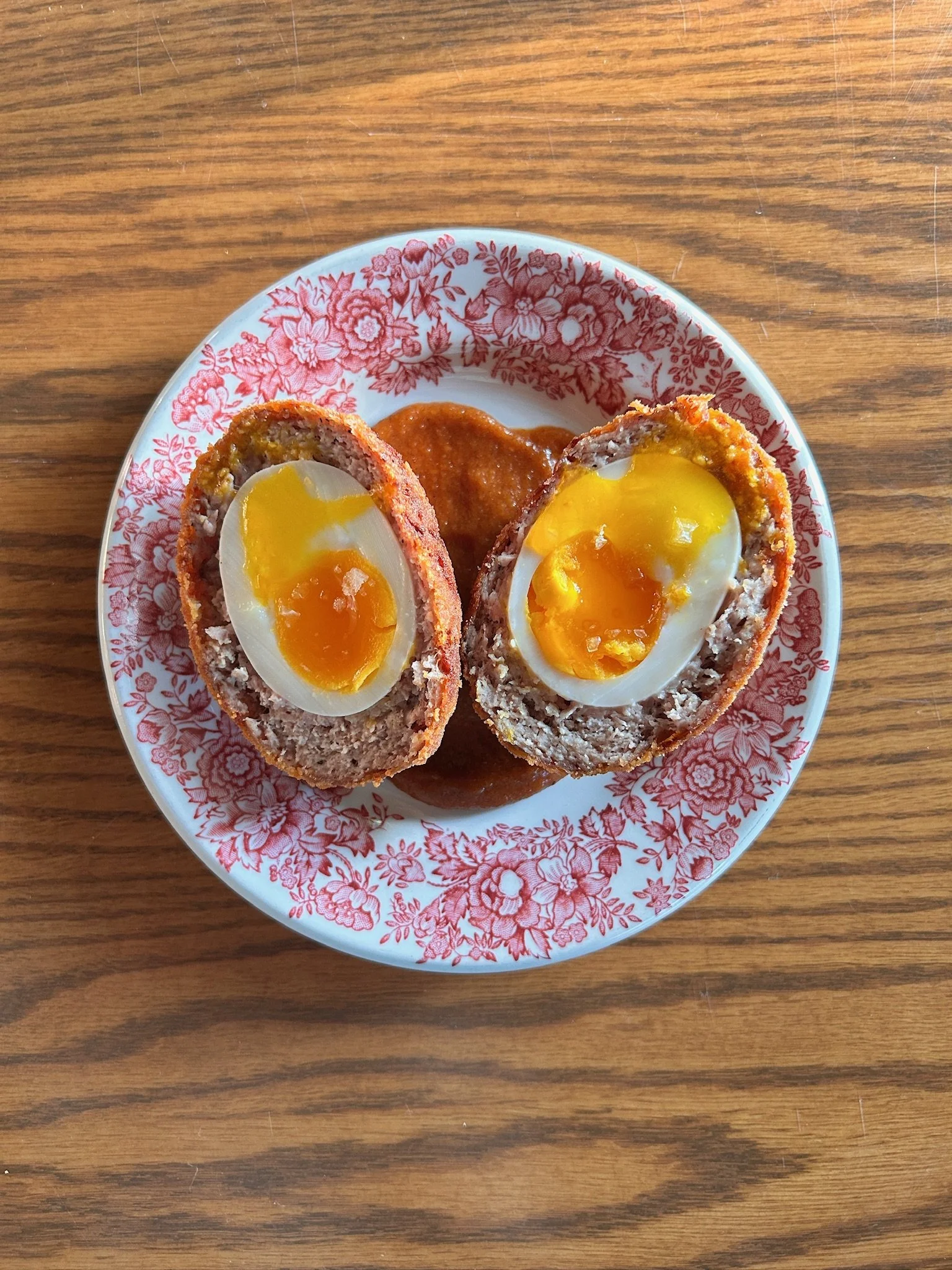 Deux œufs mollets enrobés de viande, coupés en deux, servies avec une sauce tomate dans une assiette à motif floral rouge et blanc, sur une table en bois.