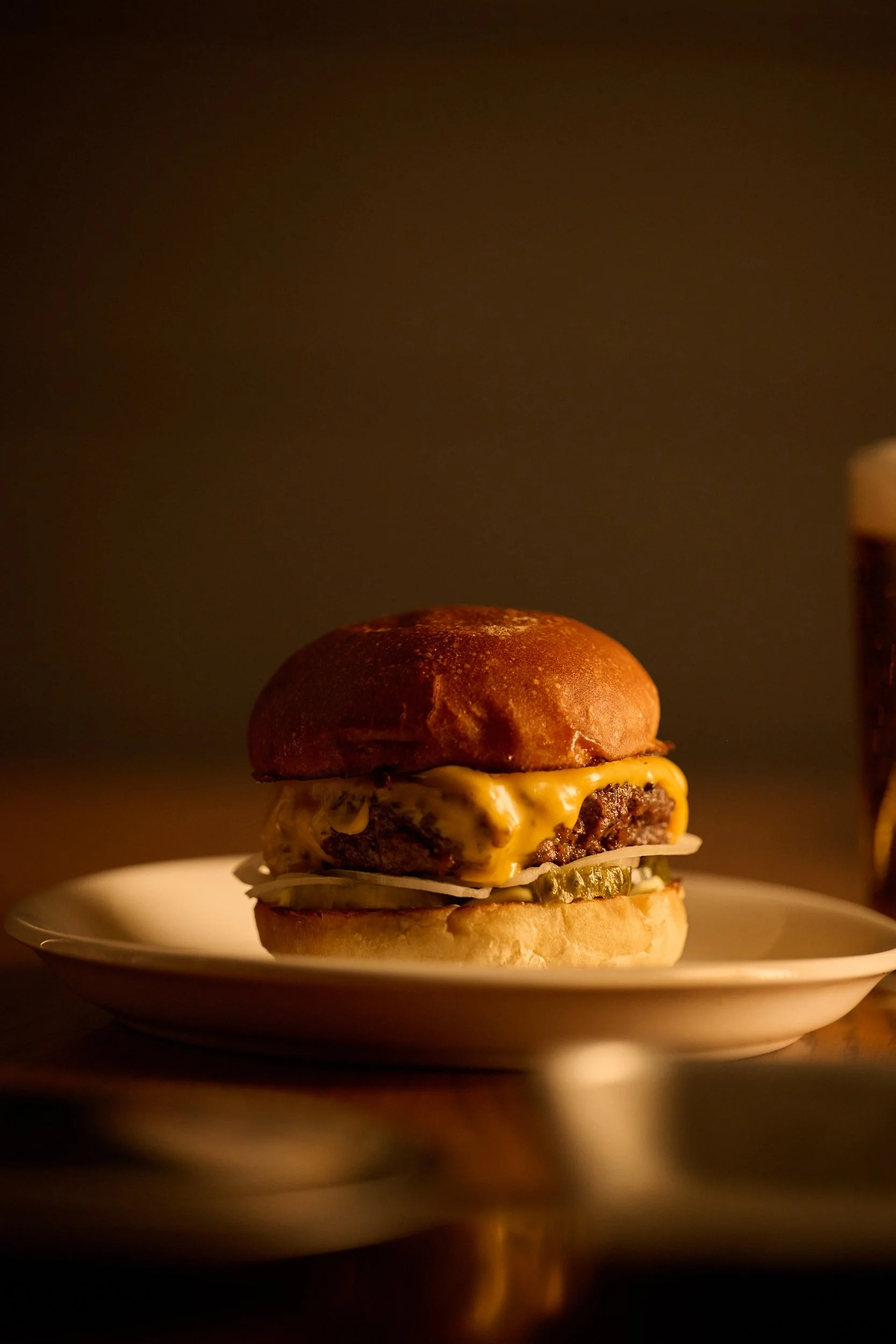 Gros plan d'un cheeseburger sur une assiette blanche à côté d'un verre de bière brune, sur fond sombre.
