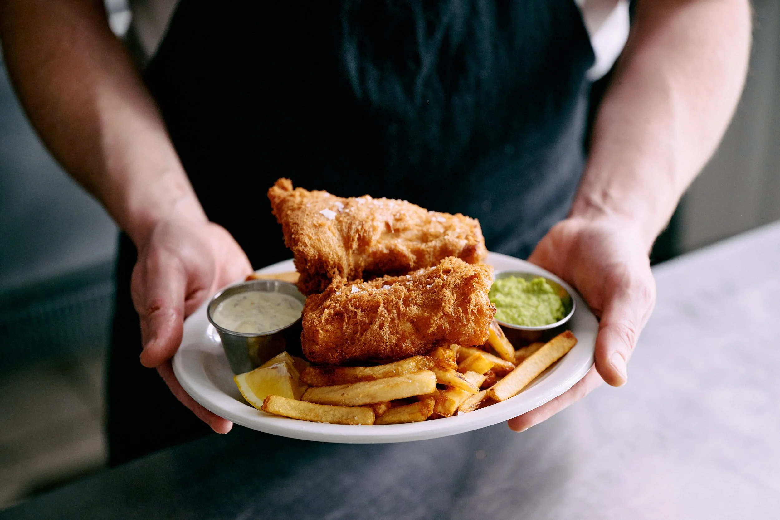 Une personne tenant une assiette blanche avec deux morceaux de poisson frit, des frites, un quartier de citron et deux petits récipients contenant de la sauce tartare et de la sauce verte.