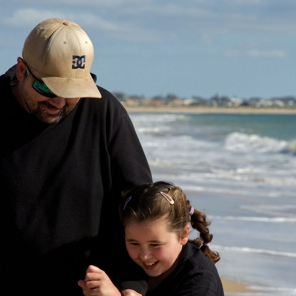 A man and a young girl laughing and playing together on the beach with the ocean waves behind them.