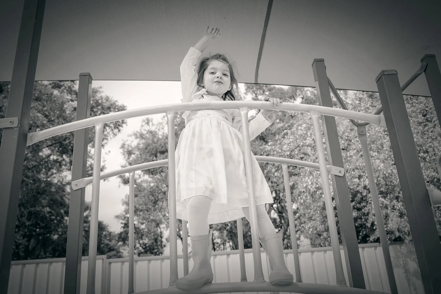 A young girl in a dress and wellington boots stands on a playground bridge with one arm in the air