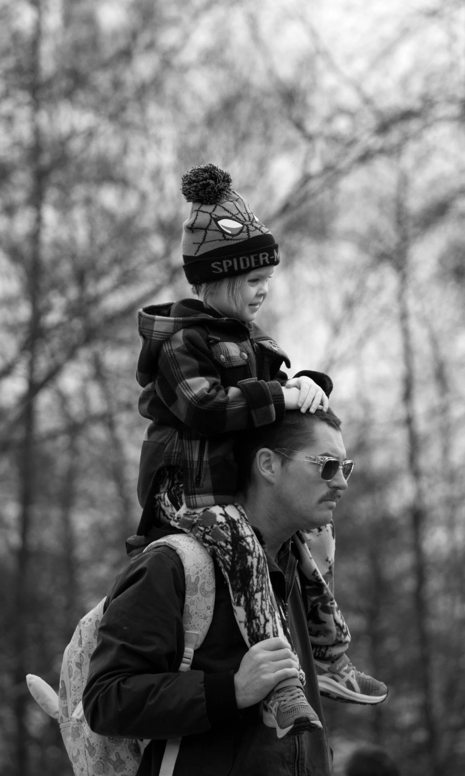 A dad carrying his young daughter on his shoulders outdoors in a wooded area, both wearing jackets and the child wearing a winter hat with a Spider-Man theme.