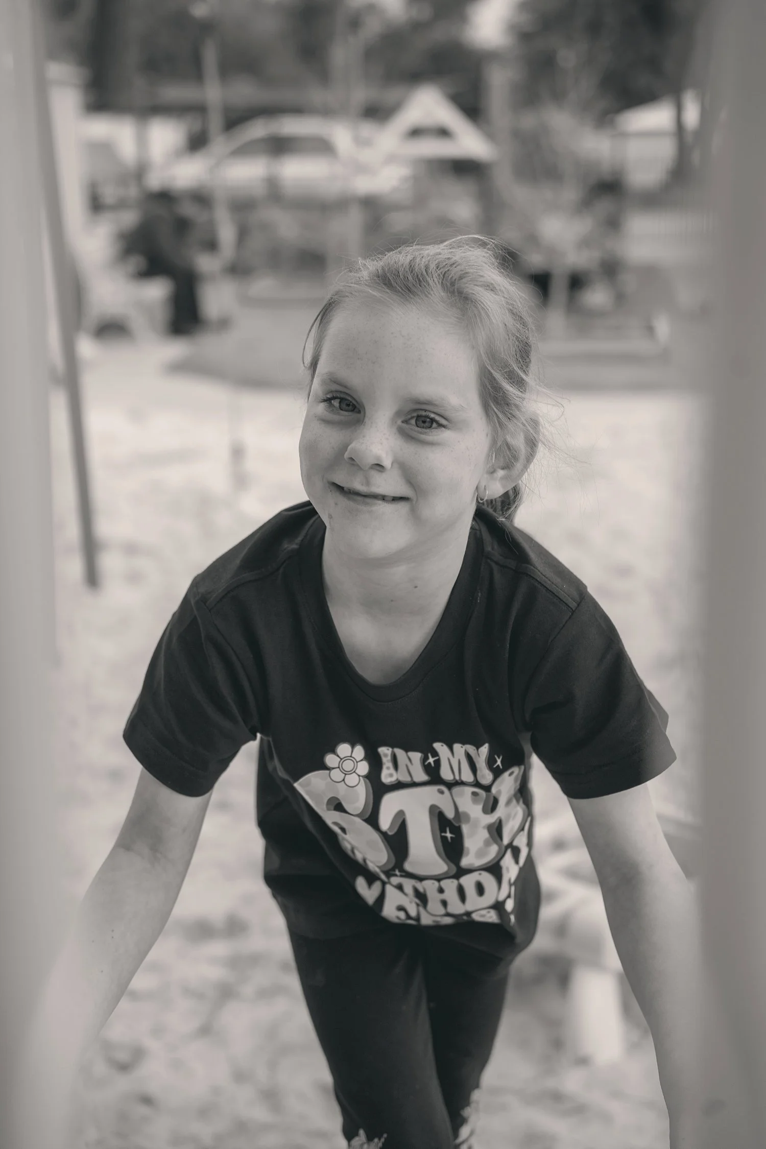 Young girl smiling at the camera, leaning forward at a playground, with playground equipment in the background.