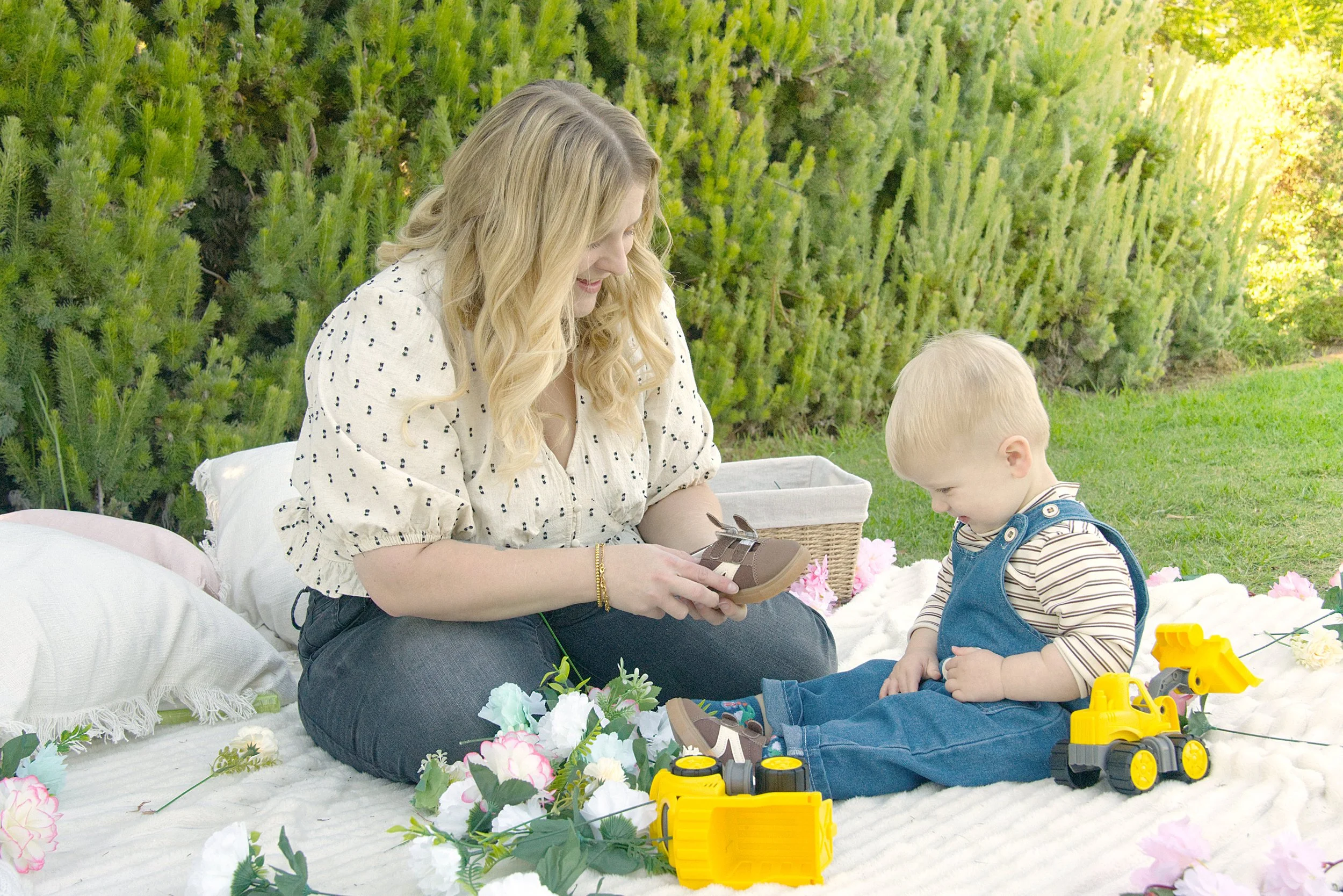 A small blonde boy  in denim dungarees looks at his feet whilst his mother smiles at him and holds a shoe.